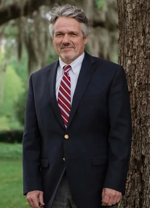 A middle-aged man with gray hair and a beard wearing a dark blazer, a white shirt, and a red striped tie, standing outdoors beside a tree with a blurred green background.