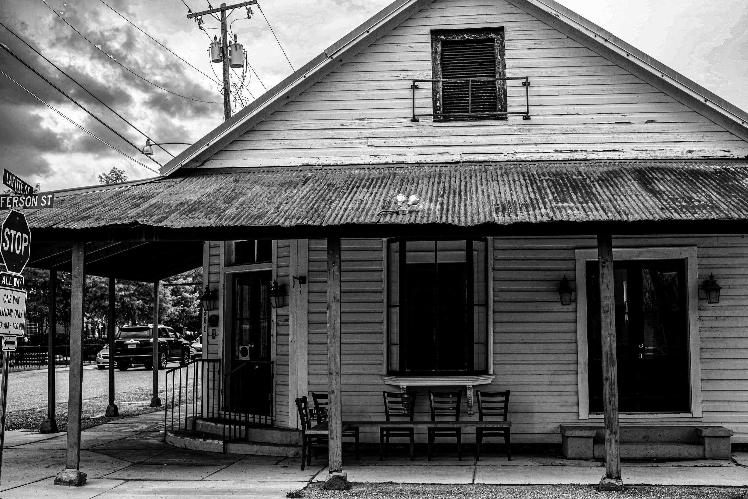 Black-and-white photo of a wooden building on a street corner, featuring a corrugated metal roof, a small balcony with a shuttered window, porch area with four chairs, street signs, and parked cars in the background.