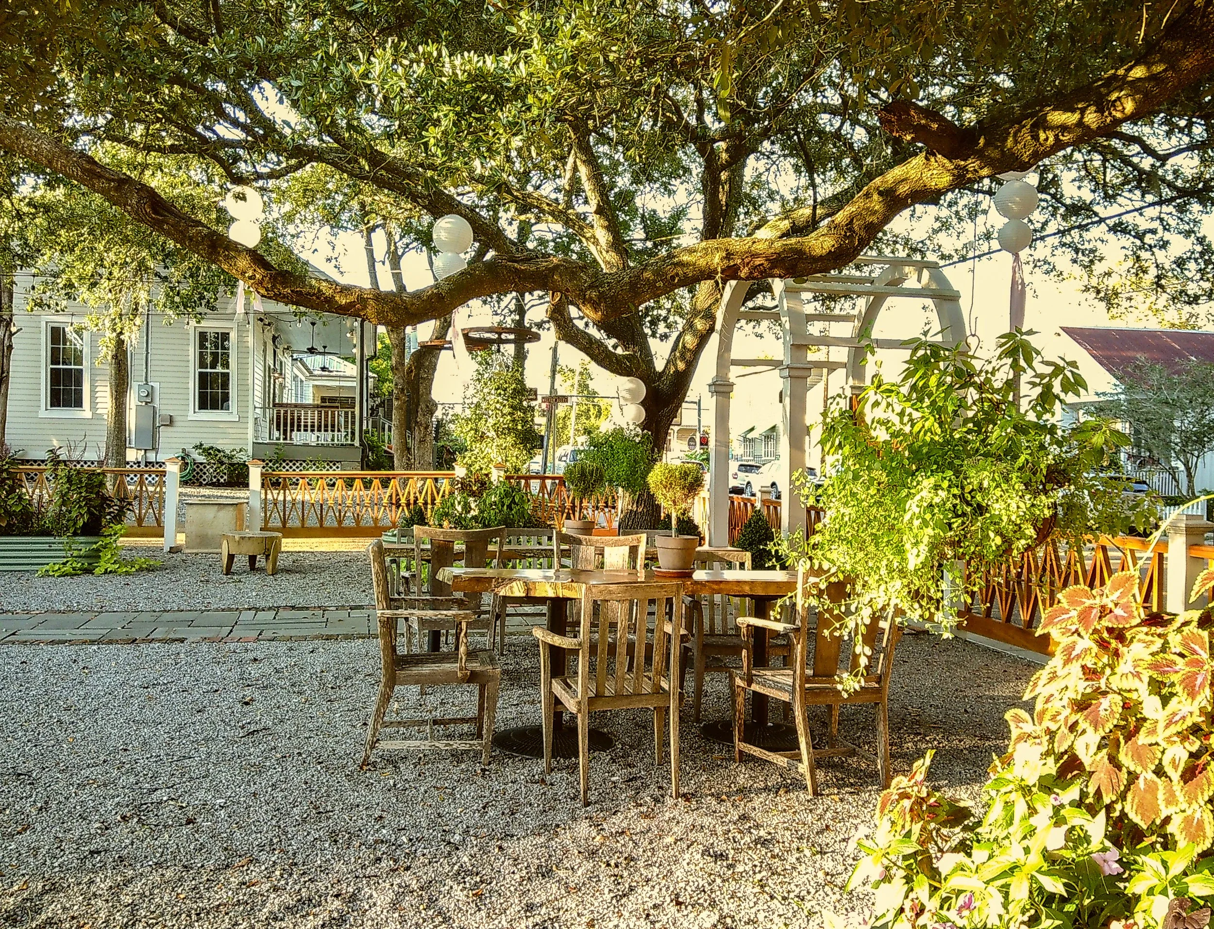 Outdoor seating area with a tree overhead, wooden chairs and tables, potted plants, a white wooden arch, and a neighboring house with white siding and a porch.
