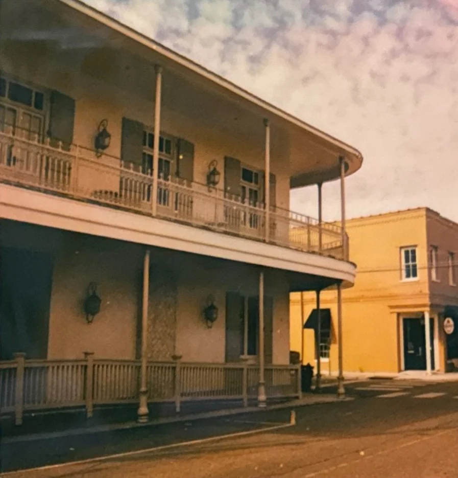 A two-story building with a balcony on the second floor and a veranda on the ground floor, featuring wooden railings, mounted lanterns, and shutters on the windows, situated on a quiet street