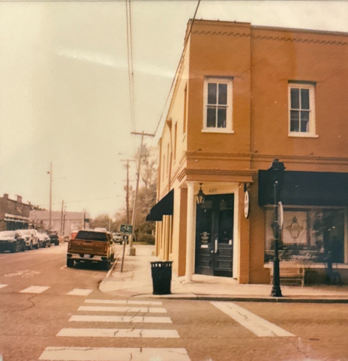 A street corner with parked cars along the side, a pedestrian crosswalk, and a two-story building with a yellow facade and black storefront on the ground floor. There are utility poles and wires, and a trash can near the sidewalk.