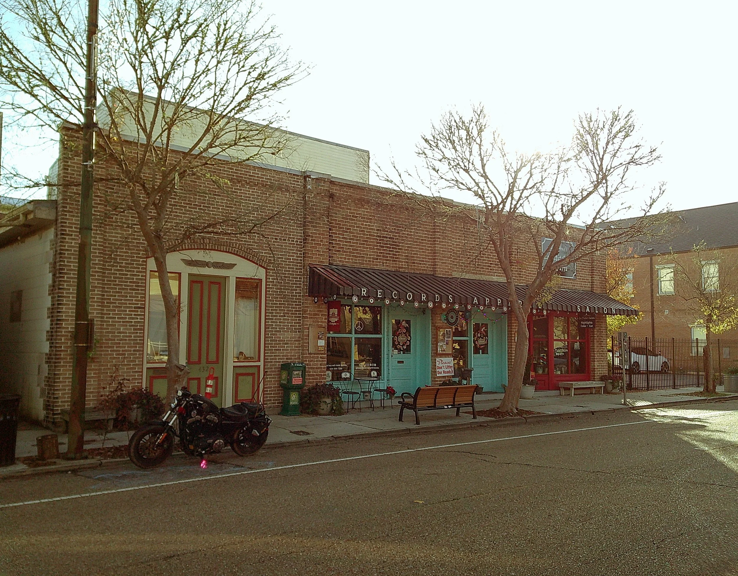 A brick building with two storefronts and a motorcycle parked outside on a street, trees with no leaves, benches, and a clear sky.