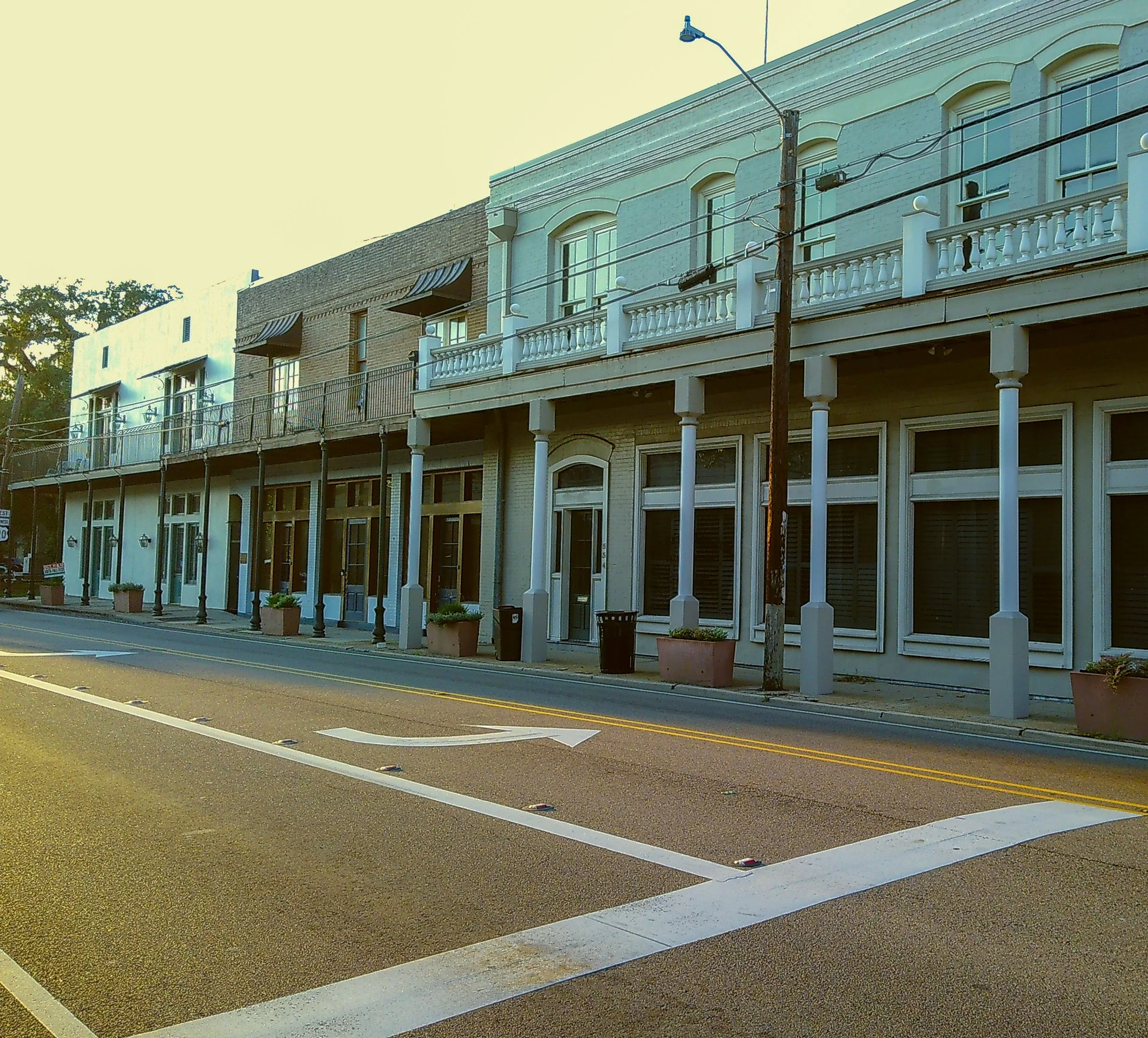 Street view of a row of commercial buildings with balconies, large windows, and potted plants, under a clear sky.