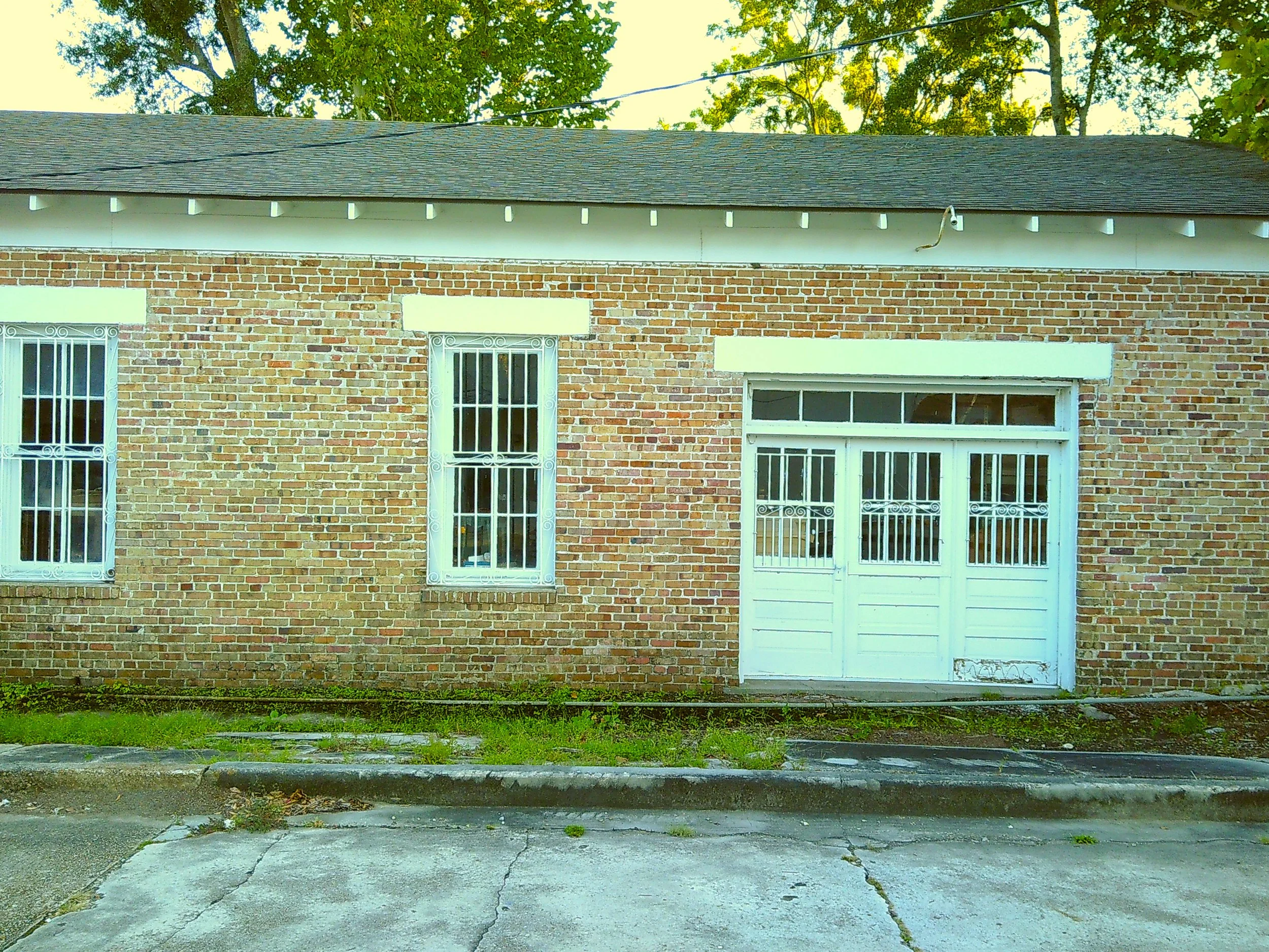 Brick building with three white metal doors and two windows with decorative white metal bars, parked cars reflected in the larger door, grassy area and curb in front, trees with green leaves in background.