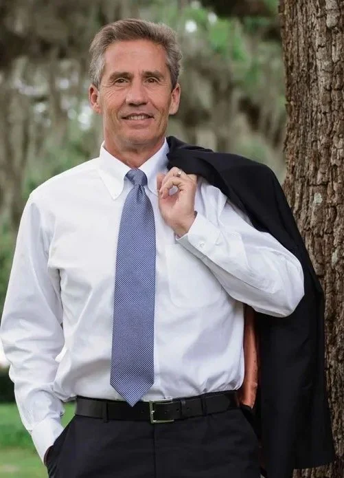A middle-aged man in a white dress shirt, gray tie, and black dress pants, standing outdoors near a tree, holding a black jacket over his shoulder, with a slight smile.