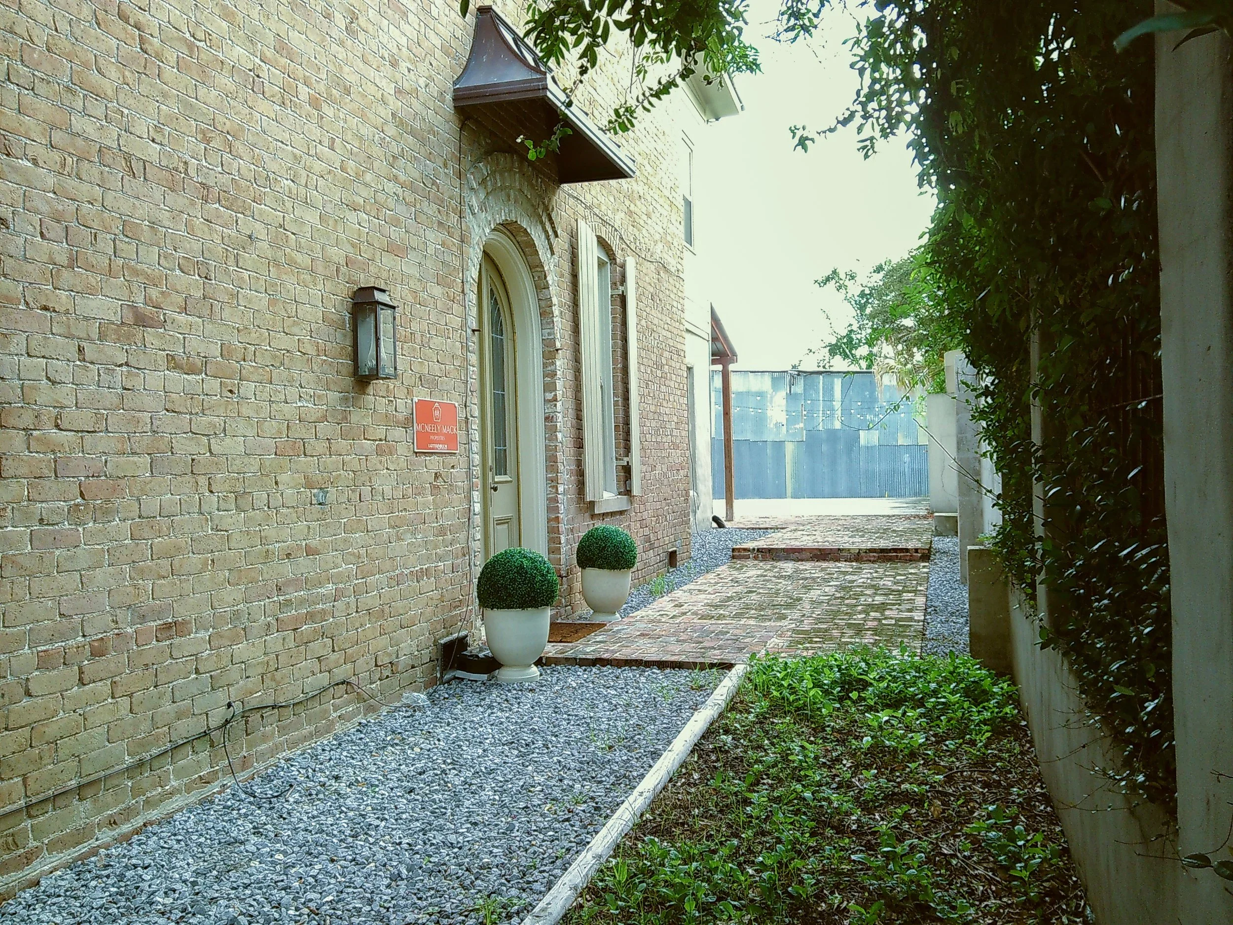 A brick house entrance with two potted topiary plants, a pathway with bricks, and a blue fence in the background.