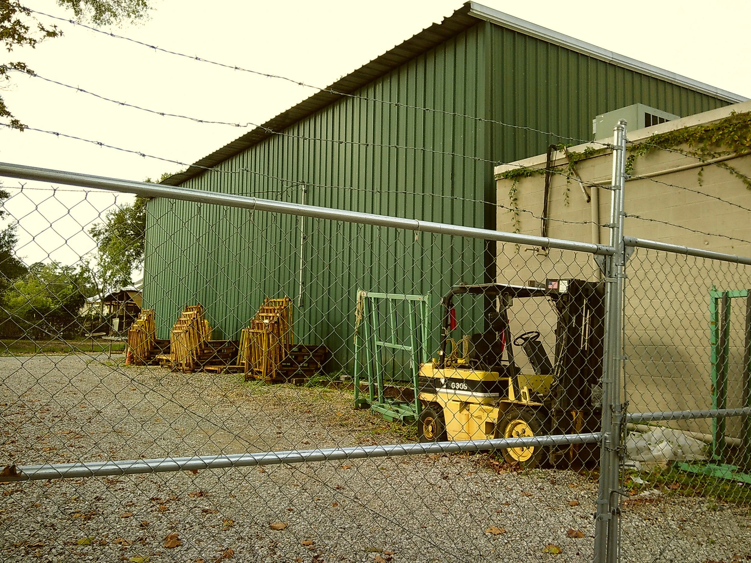 A fenced industrial area with stacked yellow ladders, a green metal building, and a small yellow forklift parked near a beige wall.