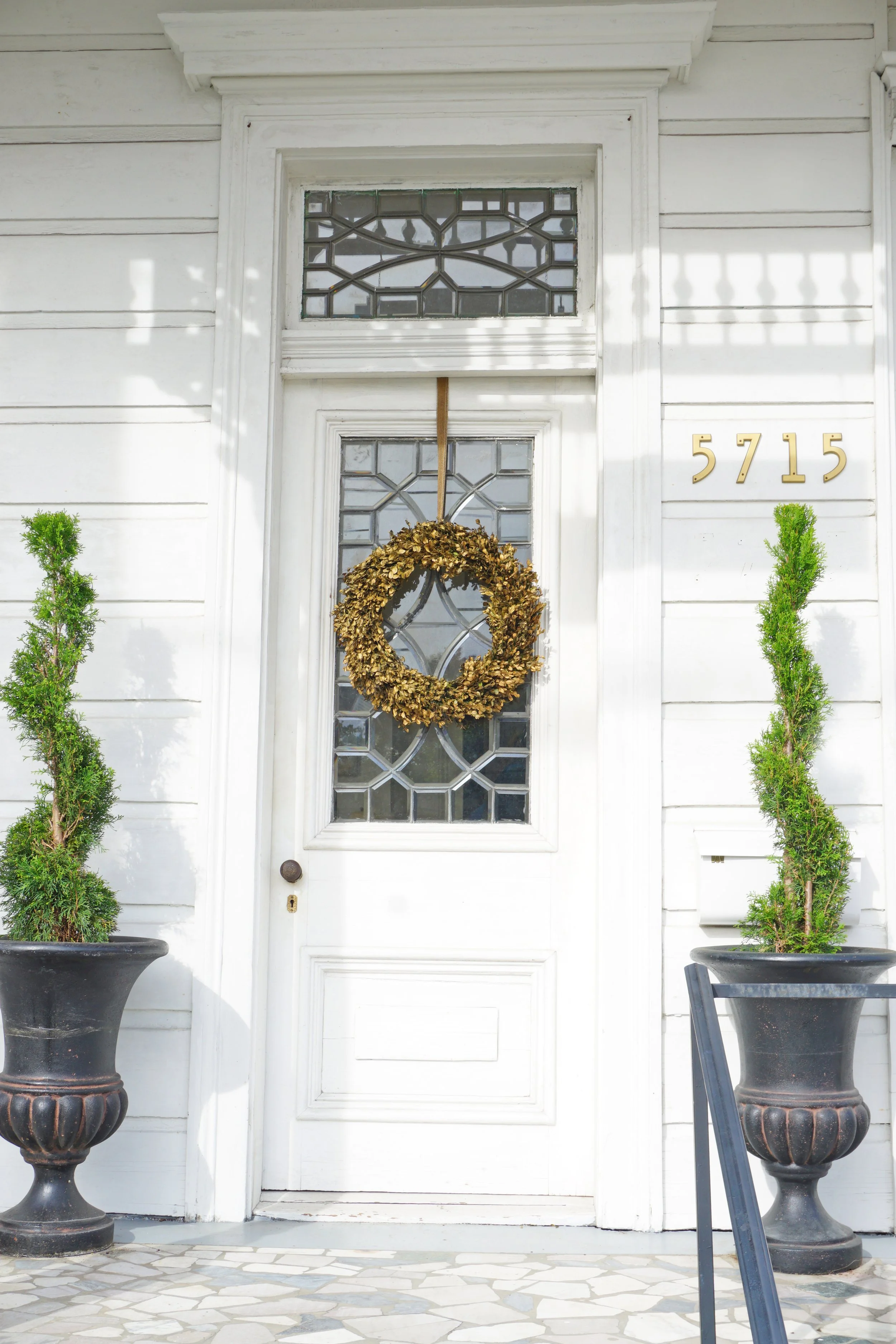 White front door with decorative glass panels, gold house number 5715, a wreath hanging on the door, and tall potted green plants on either side.