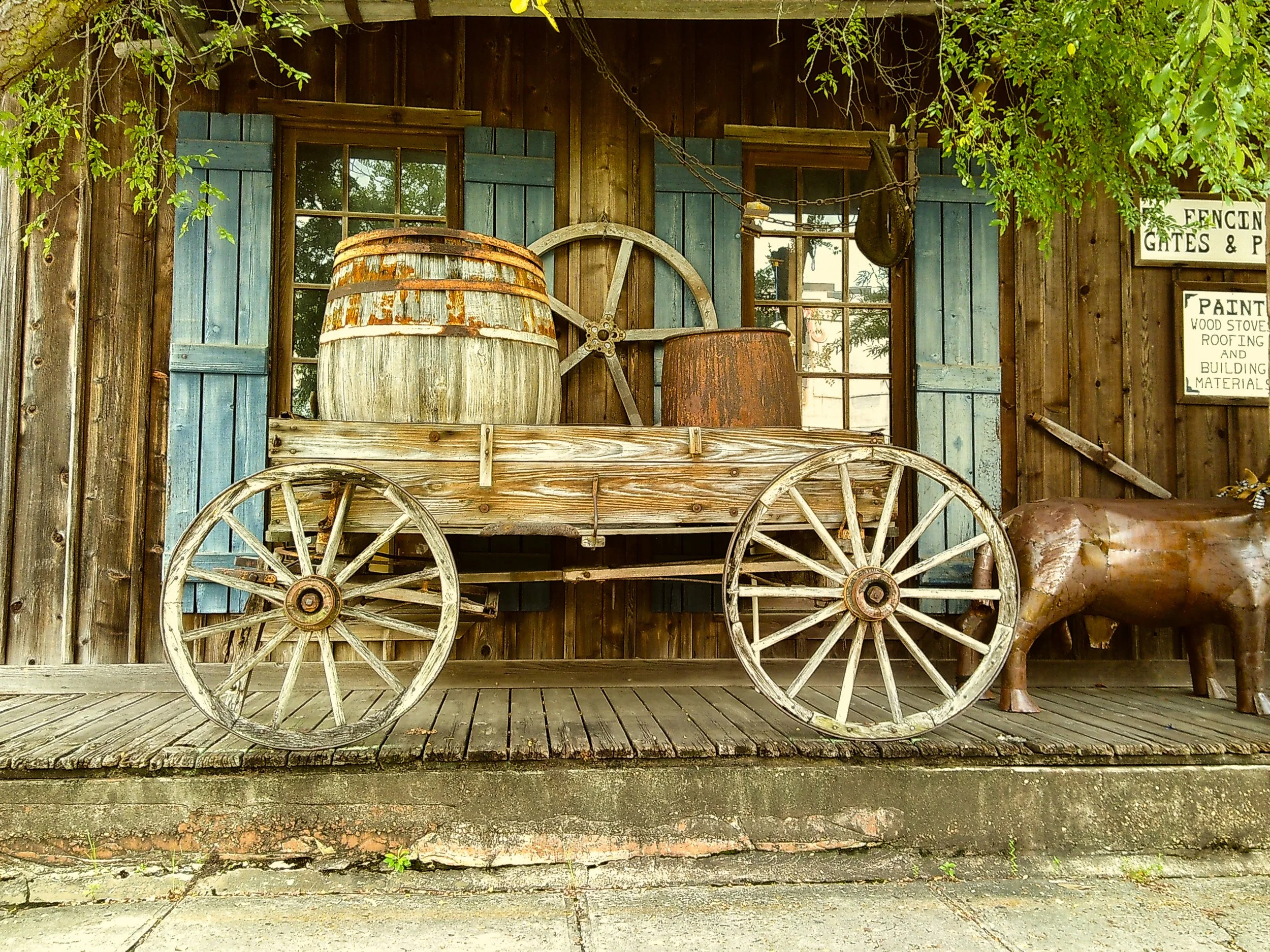 Vintage wooden cart with large wheels, barrels, and rusty metal container, set against a rustic wood building with blue shutters and signs for fencing, gates, paint, and building materials.