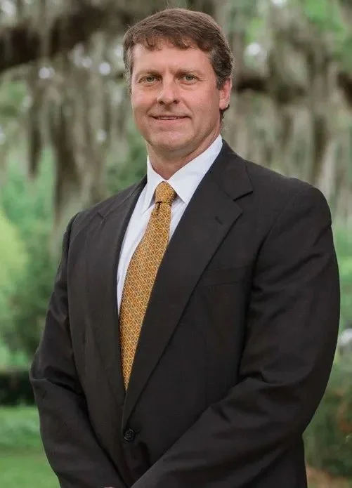 A man in a suit and tie standing outdoors with trees in the background.