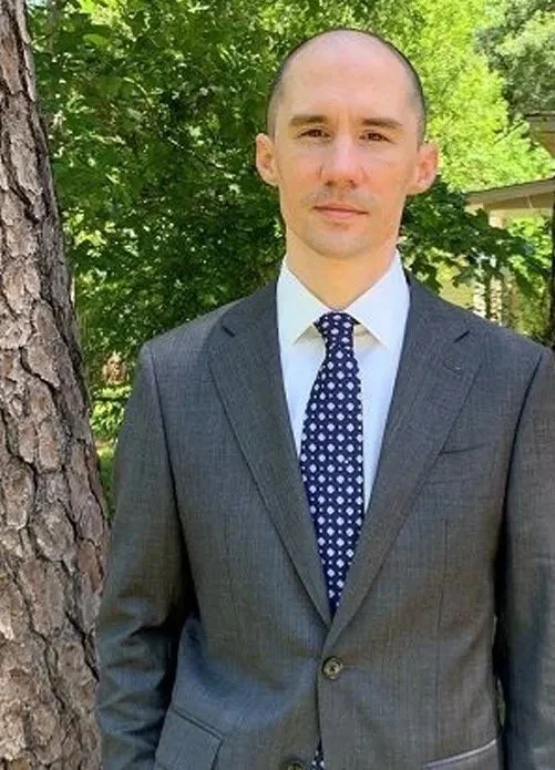 A man in a gray suit with a blue polka-dot tie standing outdoors next to a tree with green leaves.