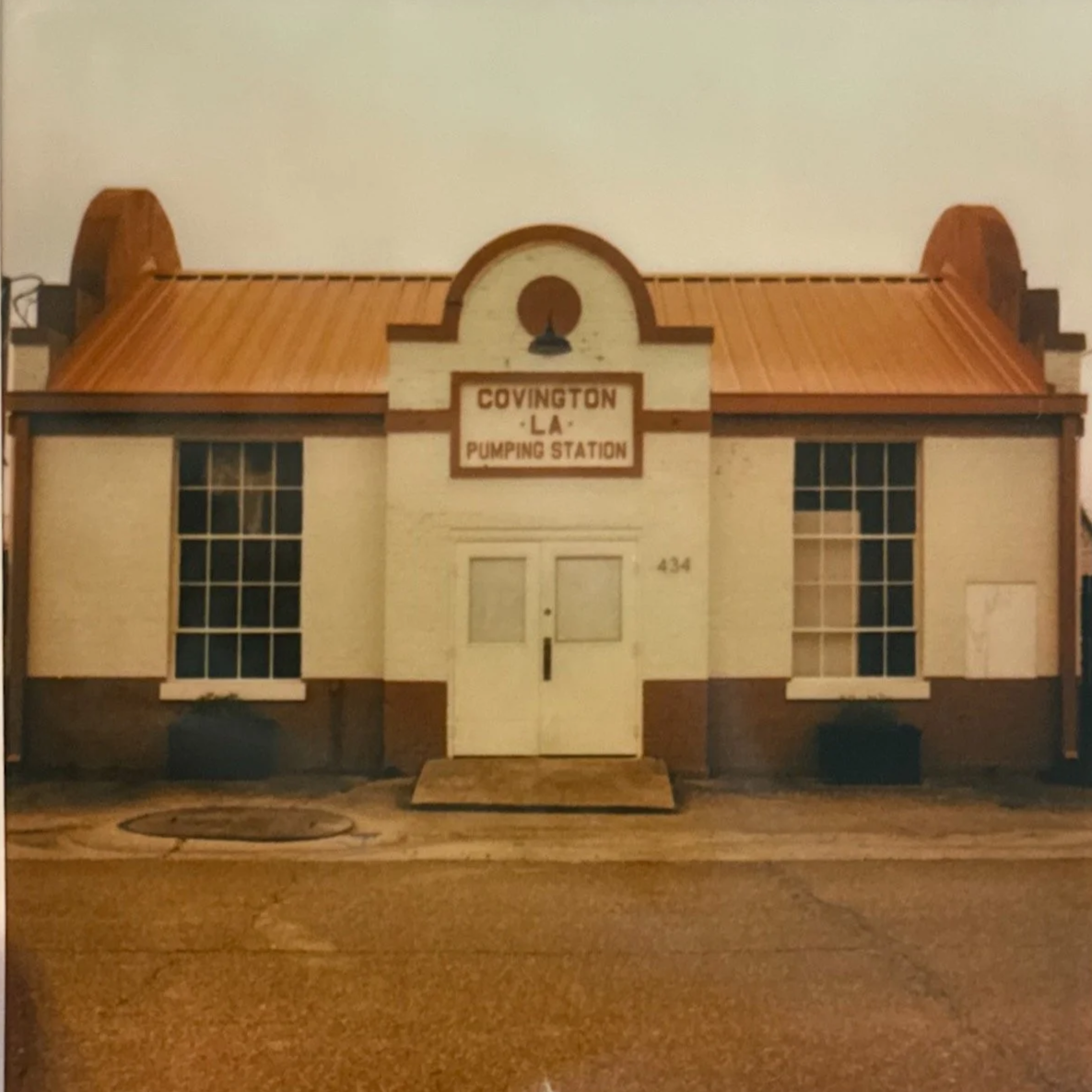 A historic building labeled 'Covington LA Pumping Station' with a symmetrical facade, two large front windows, a central double door, and a decorative sign above the entrance, featuring a curved arch design and a small light fixture.