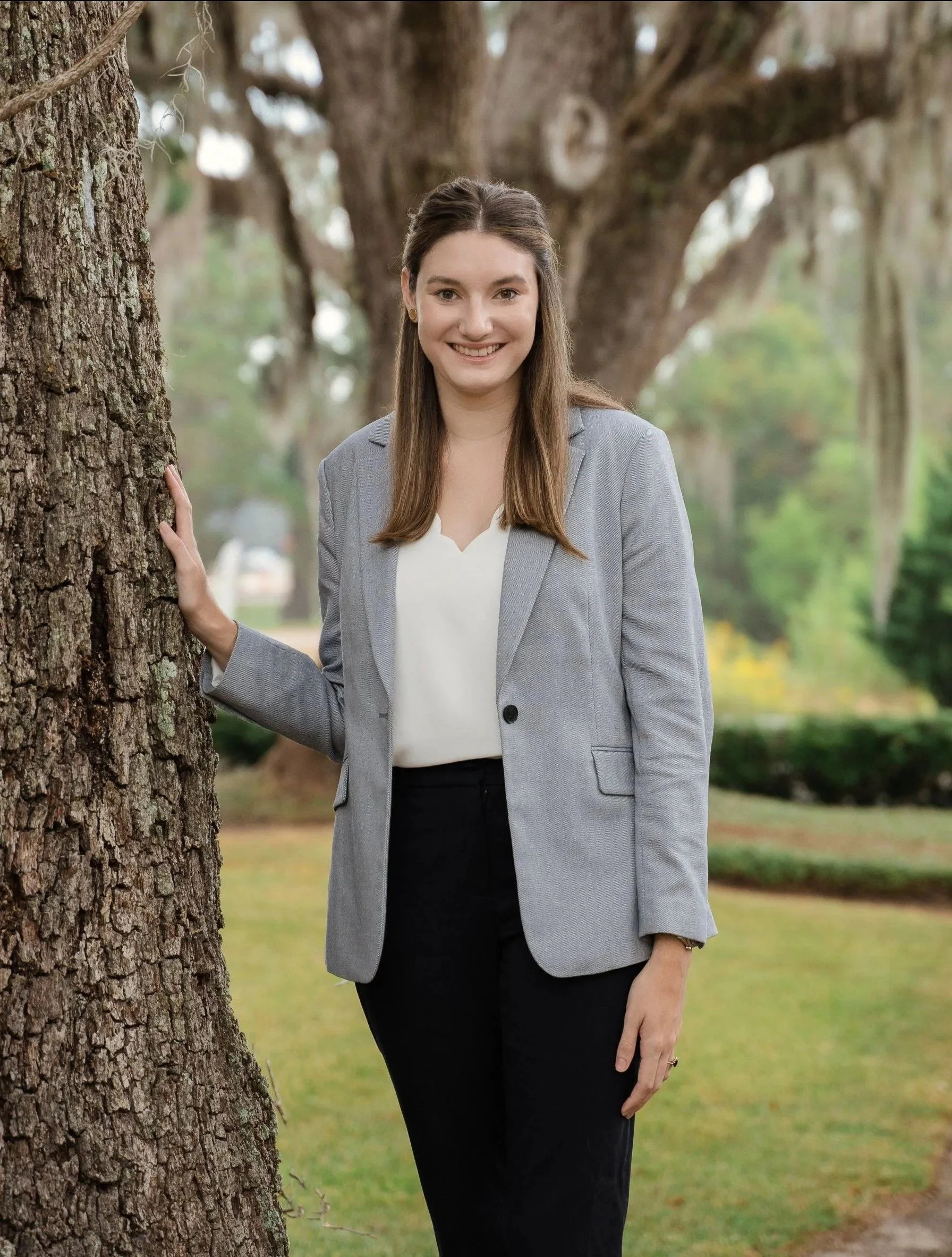 A woman with long brown hair in a gray blazer and black pants standing outdoors near a tree, smiling at the camera.