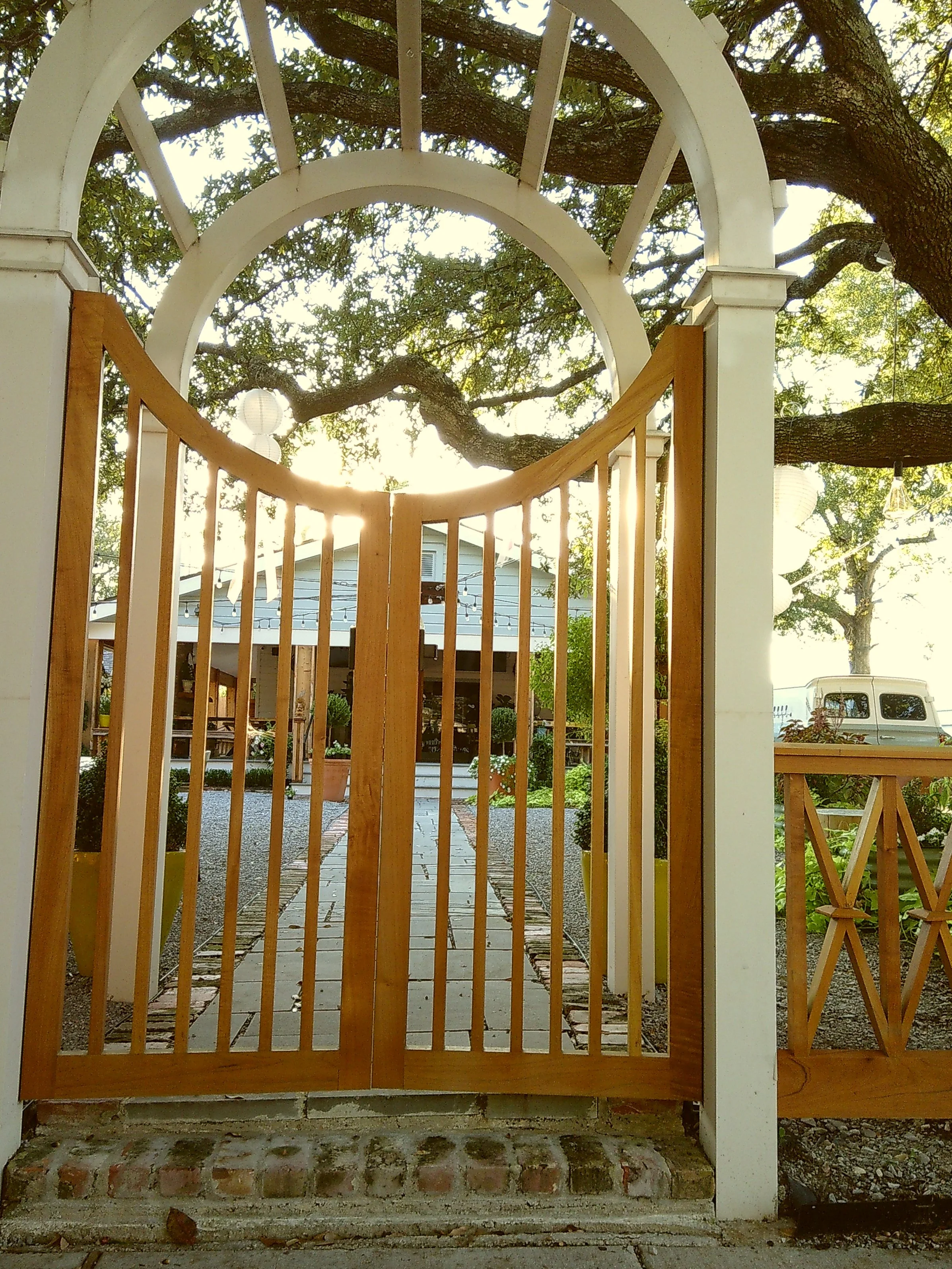 Wooden gate with arch top, open to a gravel pathway leading to a white house with a porch, surrounded by trees and decorative lanterns hanging from the branches.