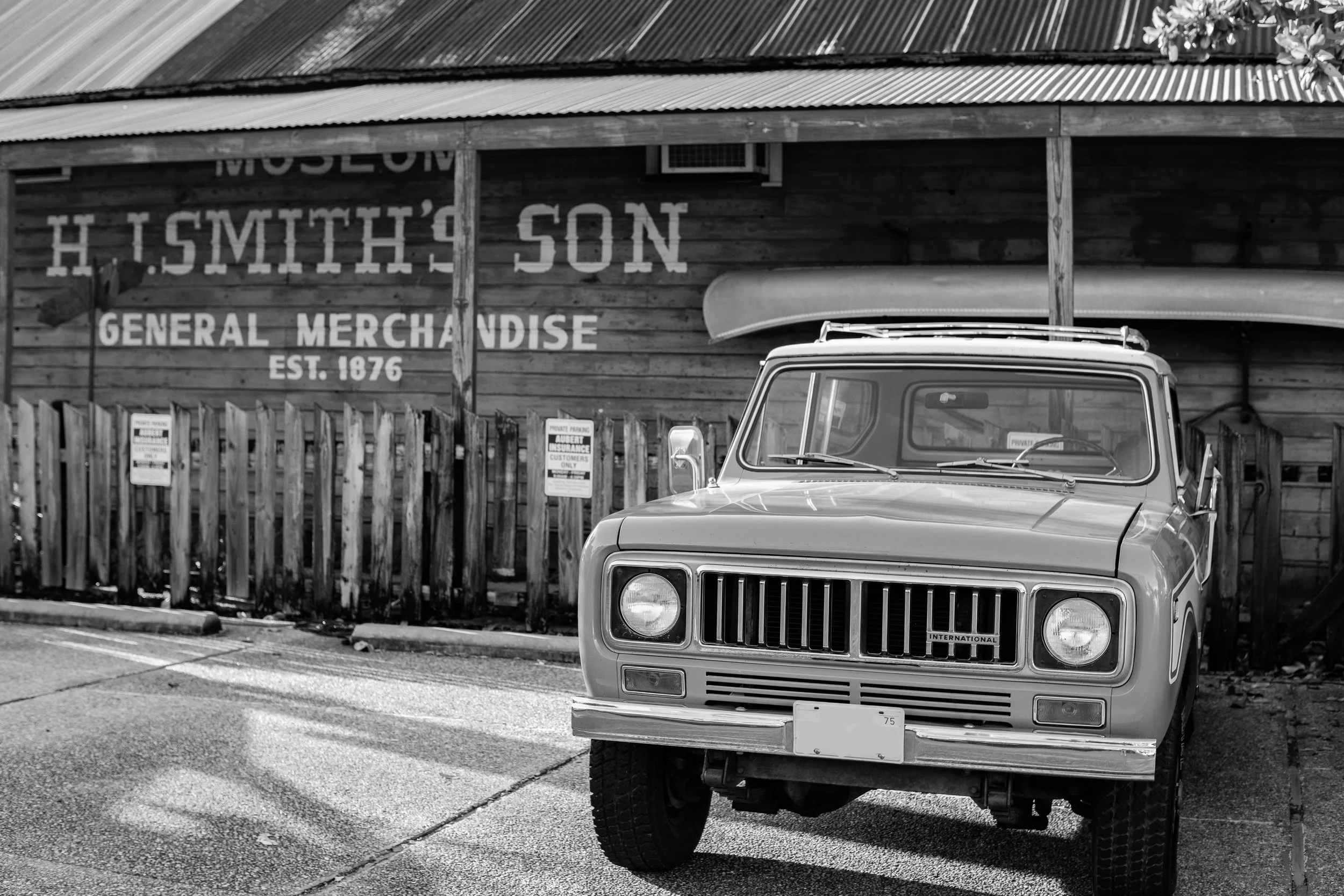 A vintage truck parked in front of a wooden building with sign reading "H. T. Smith's Son General Merchandise Est. 1876." A surfboard is mounted on the roof of the truck.
