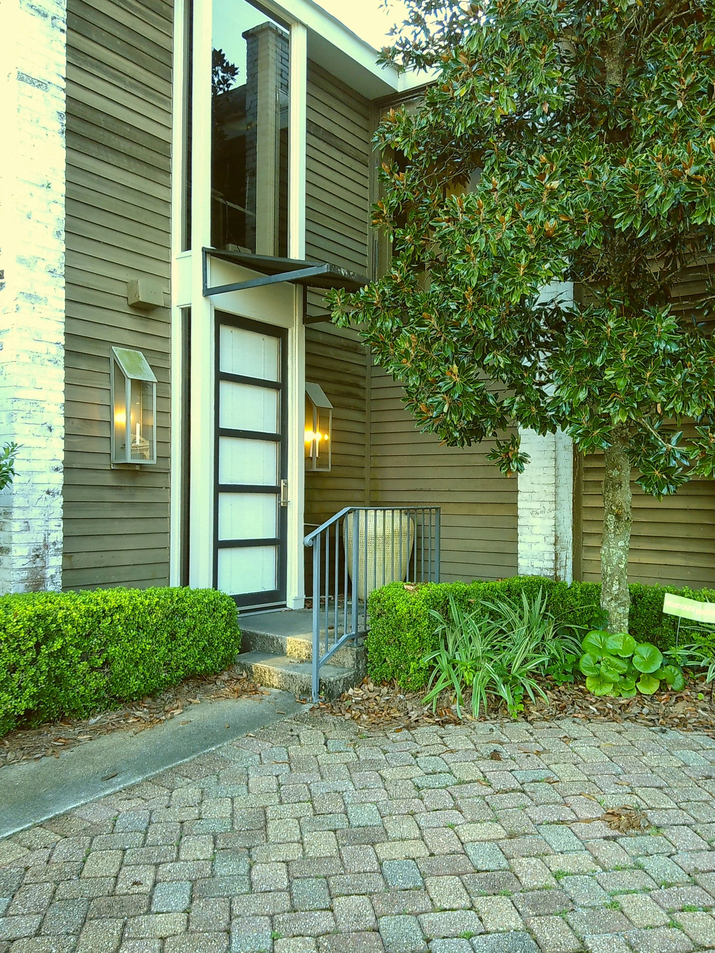 The front entrance of a modern residential building with a white door, black framing, and outdoor lighting fixtures, surrounded by trimmed bushes, a tree, and potted plants on a paved stone pathway.