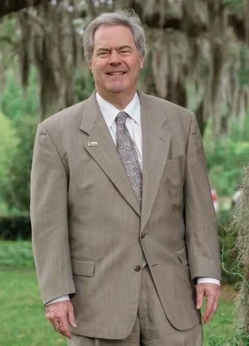 A smiling man wearing a beige suit with a tie, standing outdoors in a green park with trees in the background.
