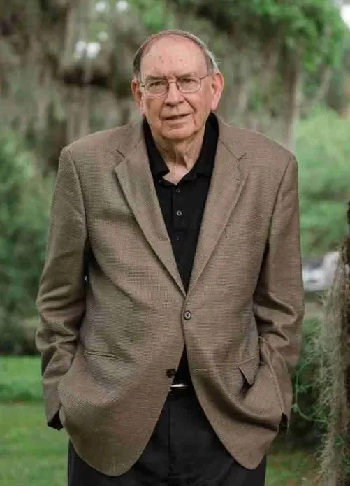 An elderly man with glasses wearing a brown blazer and black shirt standing outdoors in a green park.