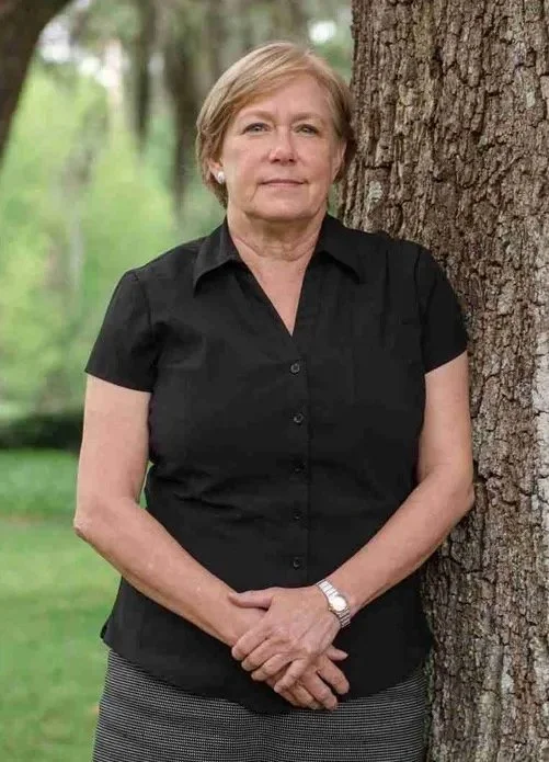 An older woman with short blonde hair standing next to a large tree outdoors. She is wearing a black short-sleeved shirt, a patterned skirt, and a watch, with her hands clasped in front of her.