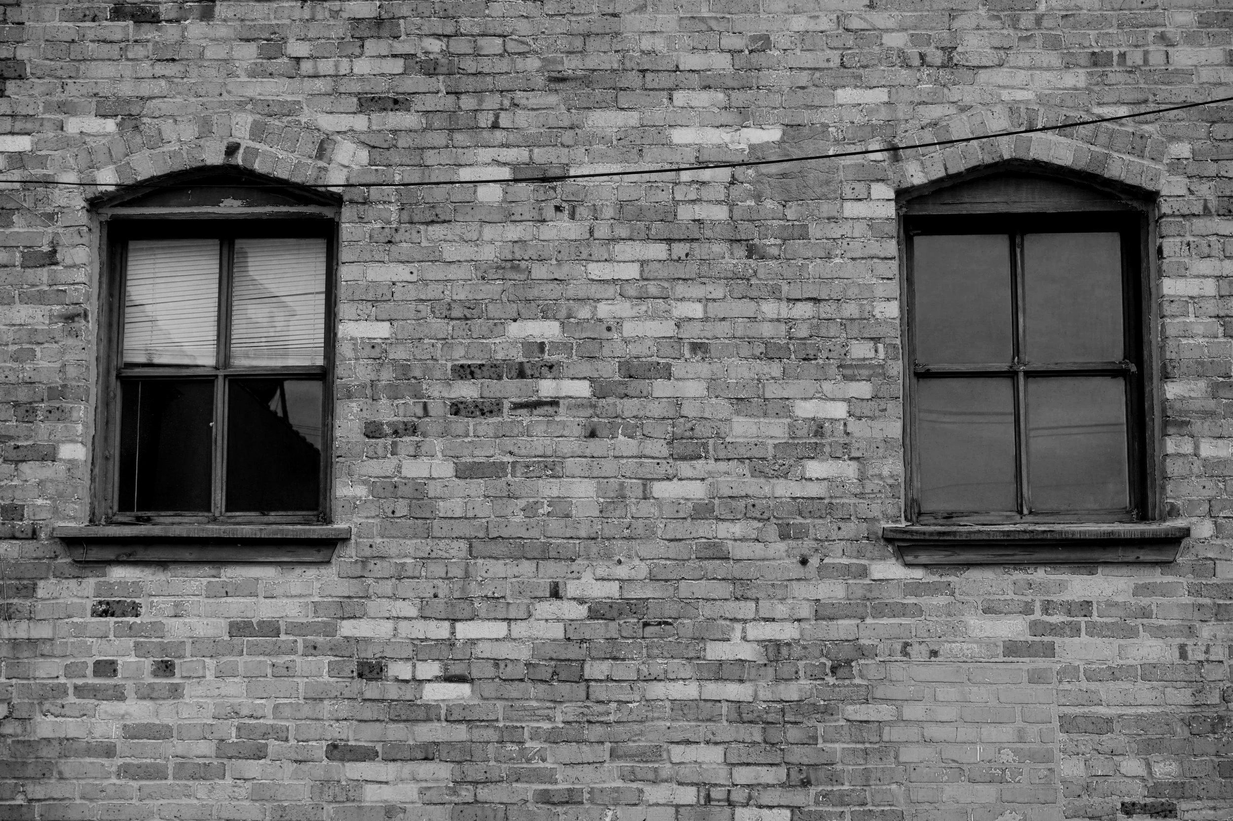 Black and white photograph of an old brick building facade with two windows, one on the left and one on the right, both with wooden frames and windowpanes.