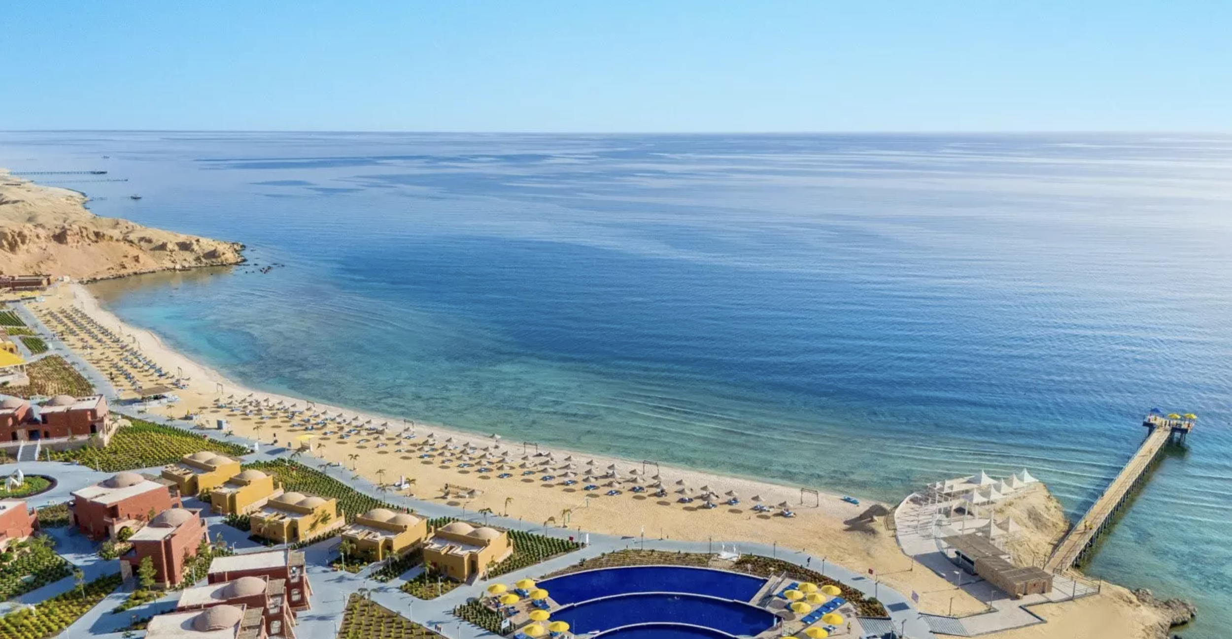 Spiaggia con ombrelloni e sdraio, mare calmo e acqua cristallina, palazzi e strutture ricreative sulla spiaggia, un molo che si estende in mare sulla destra