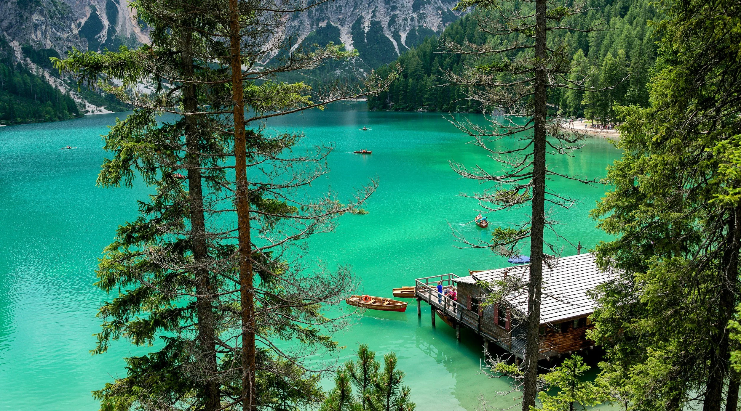 Lago turchese circondato da foresta e montagne, con un pontile in legno e alcune barche parcheggiate, visibile attraverso gli alberi.