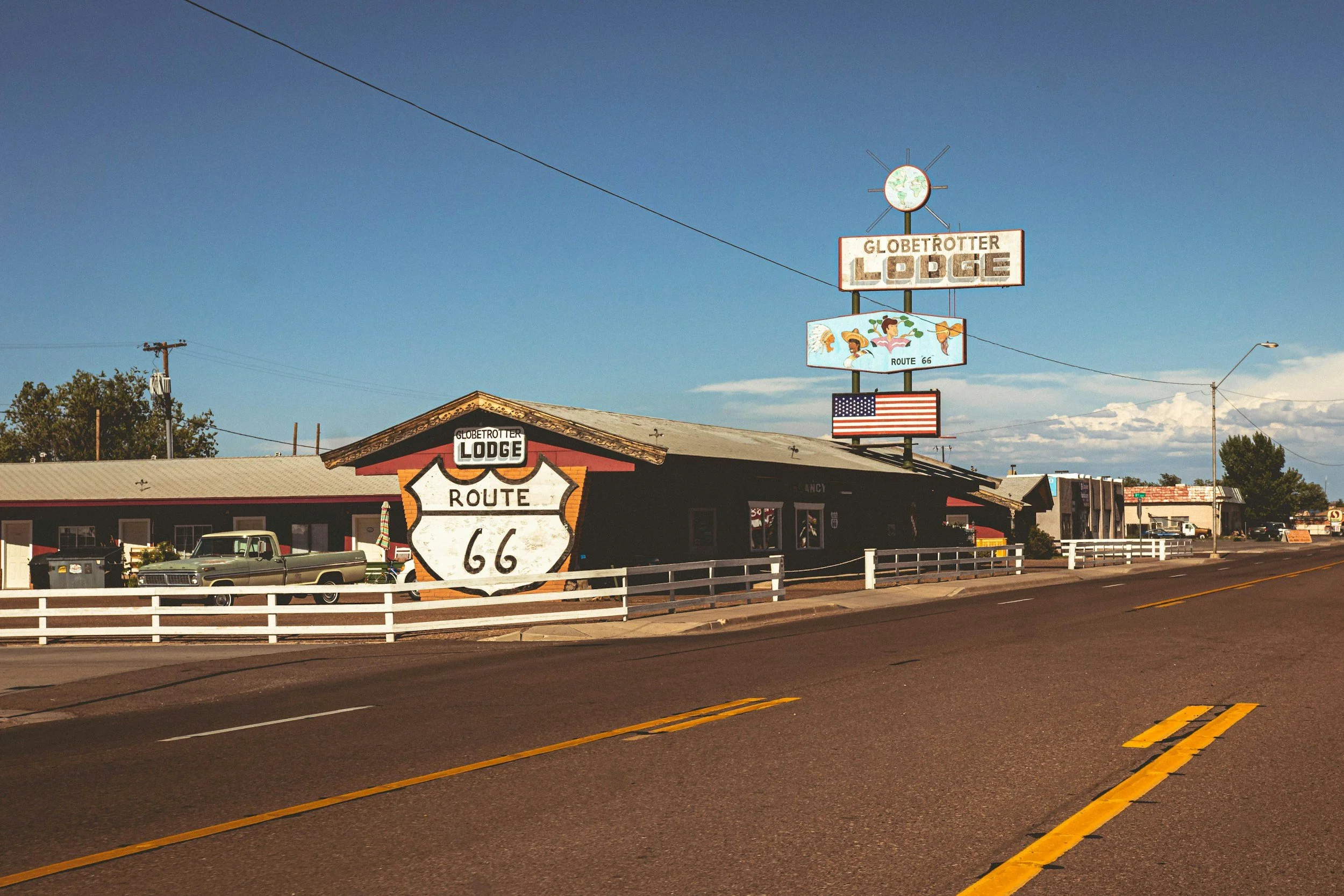 Struttura in legno con segnali Route 66, visto di fronte, con auto vintage parcheggiate davanti, cielo azzurro e luci stradali.