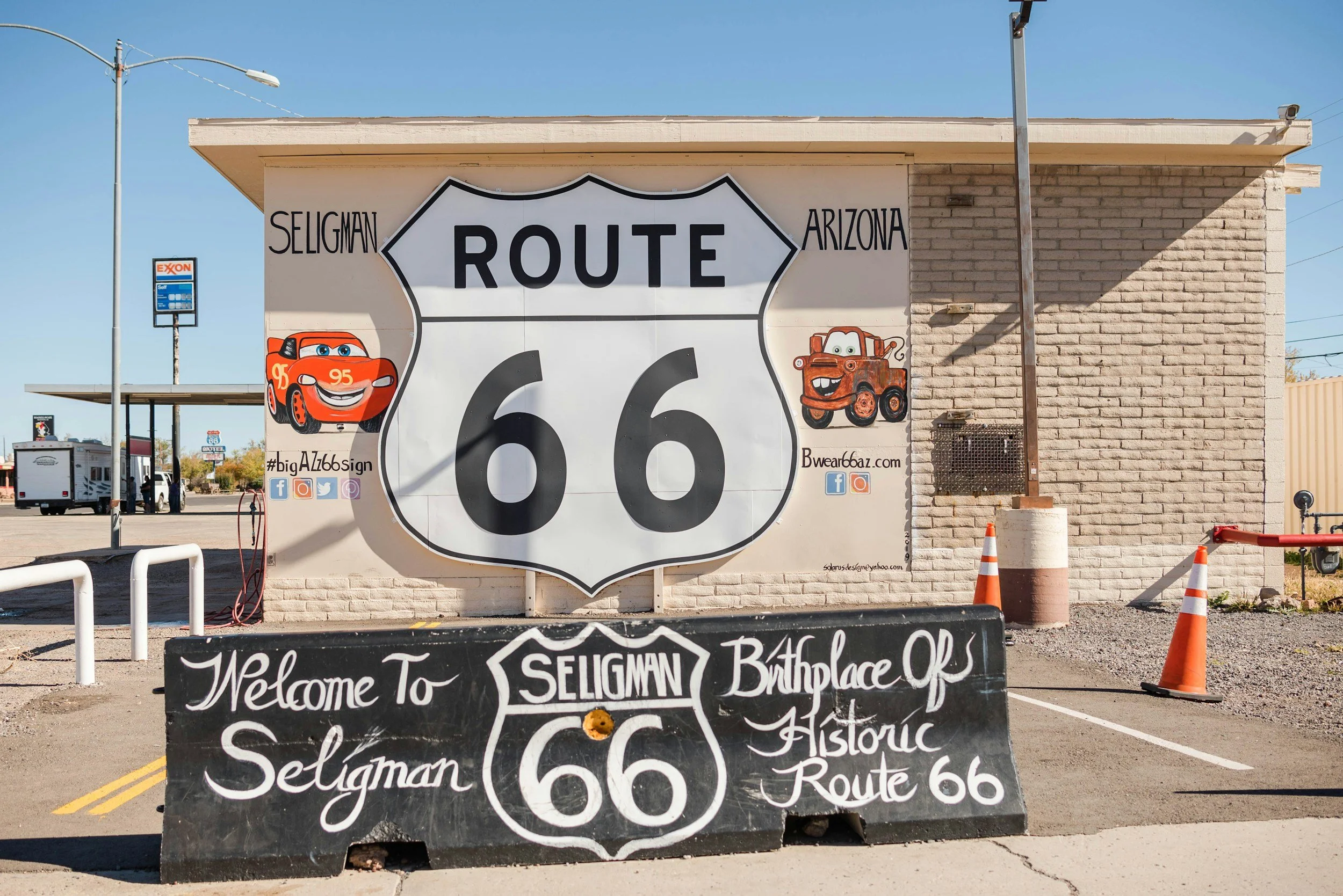 Murale con cartello di Route 66, con le parole Seligman, Arizona, e due personaggi di cartone animato di veicoli, uno rosso e uno marrone. Cartello scritto a mano: Welcome to Seligman, birthplace of historic Route 66.