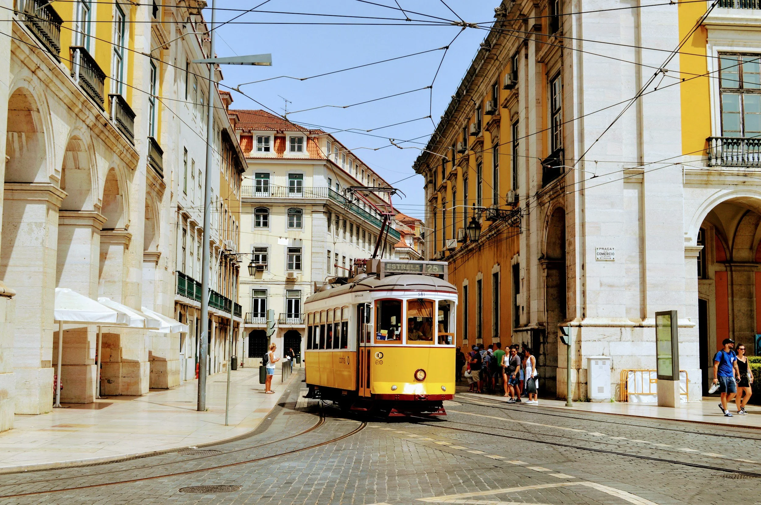 Tram giallo che attraversa una strada nel centro storico di Lisbona, tra edifici storici e persone che camminano.