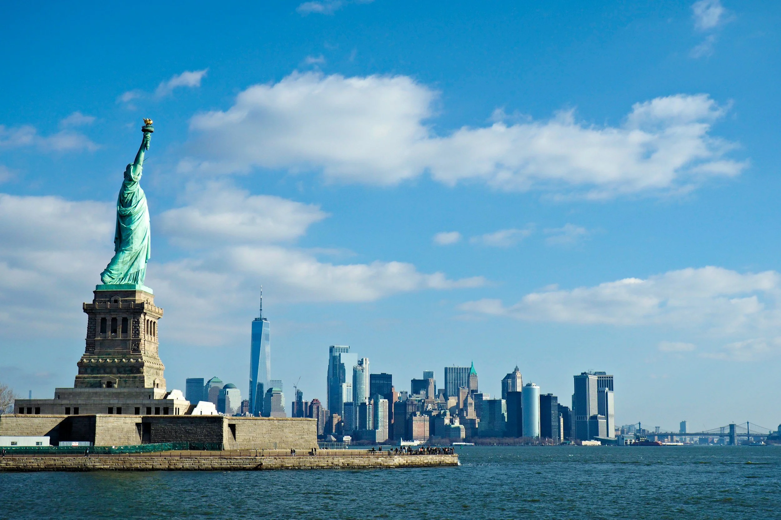 Statua della Libertà vista dal porto di New York con lo skyline di Manhattan e il ponte di Brooklyn in background, cielo blu con qualche nuvola.