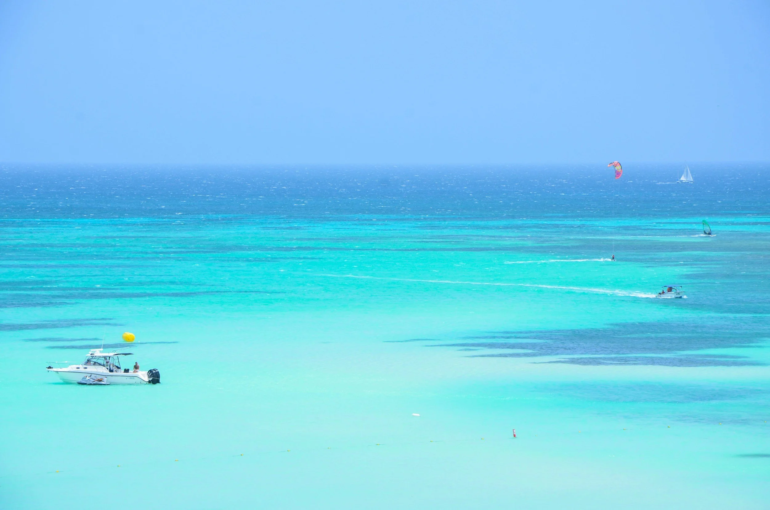 Mare turchese con barche a vela e motoscafi, cielo azzurro, e alcune persone che praticano sport acquatici come il kitesurf.