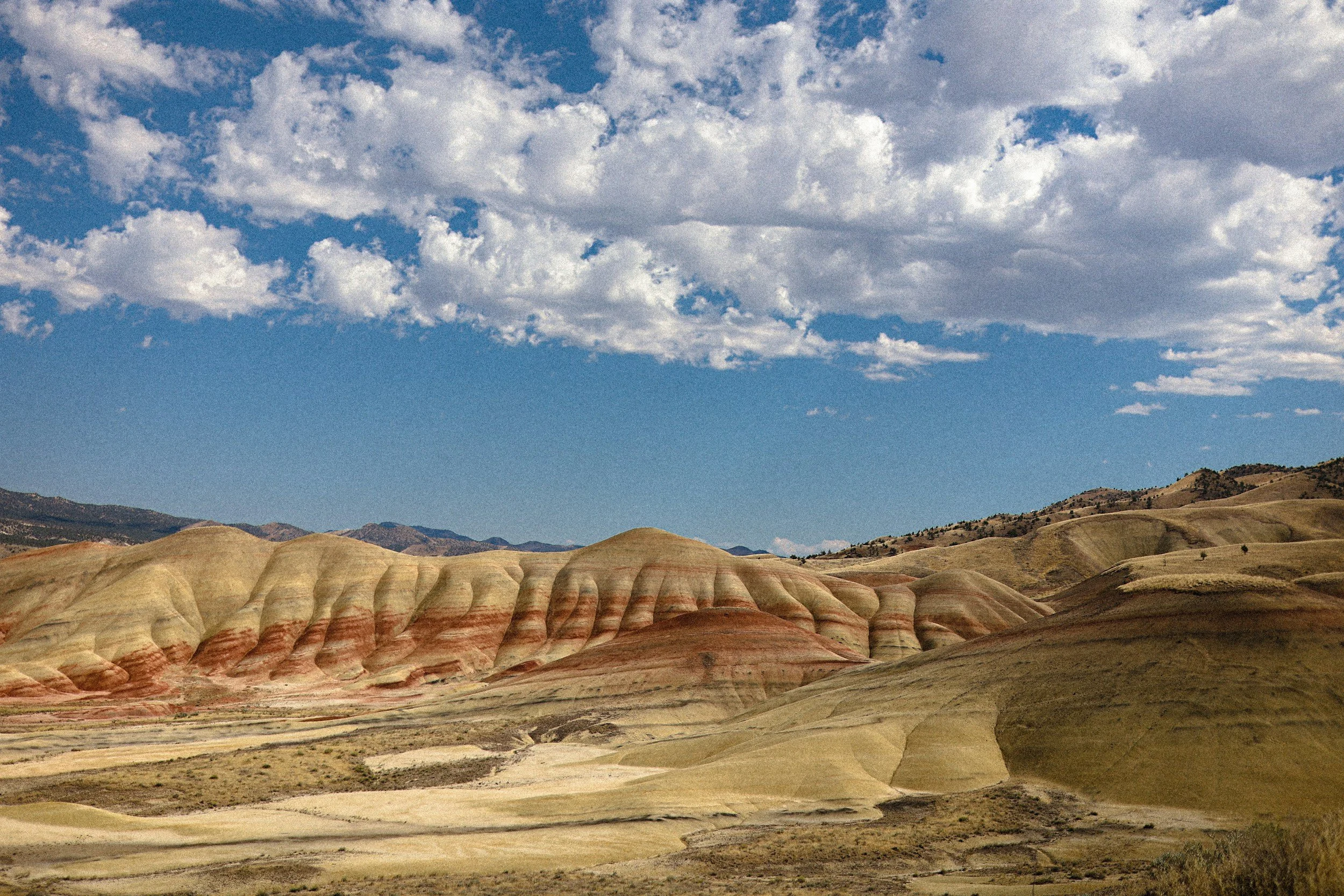 Paesaggio naturale con colline ondulate e striature di diversi colori, cielo nuvoloso con nuvole bianche e cielo blu.