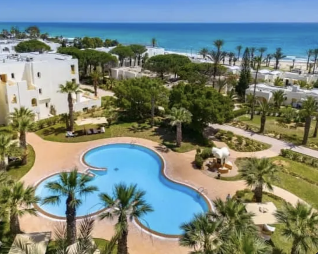 Vista di una piscina a forma di nuvola in un resort vicino alla spiaggia con ombrelloni, palme e case bianche sullo sfondo del mare blu.