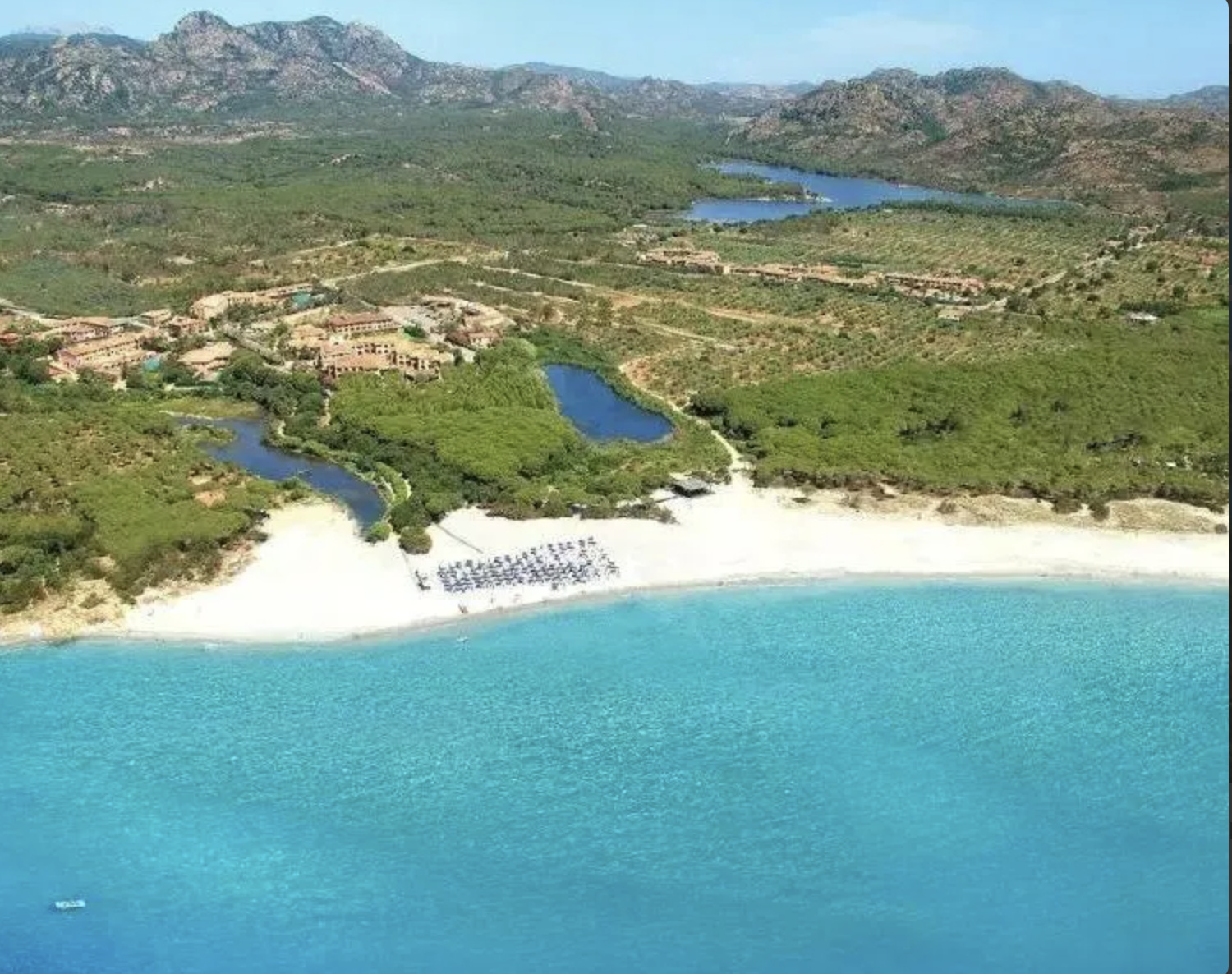 Paesaggio costiero con spiaggia di sabbia bianca, mare turchese, vegetazione verde e laghi nel contesto di un ambiente naturale con montagne sullo sfondo.