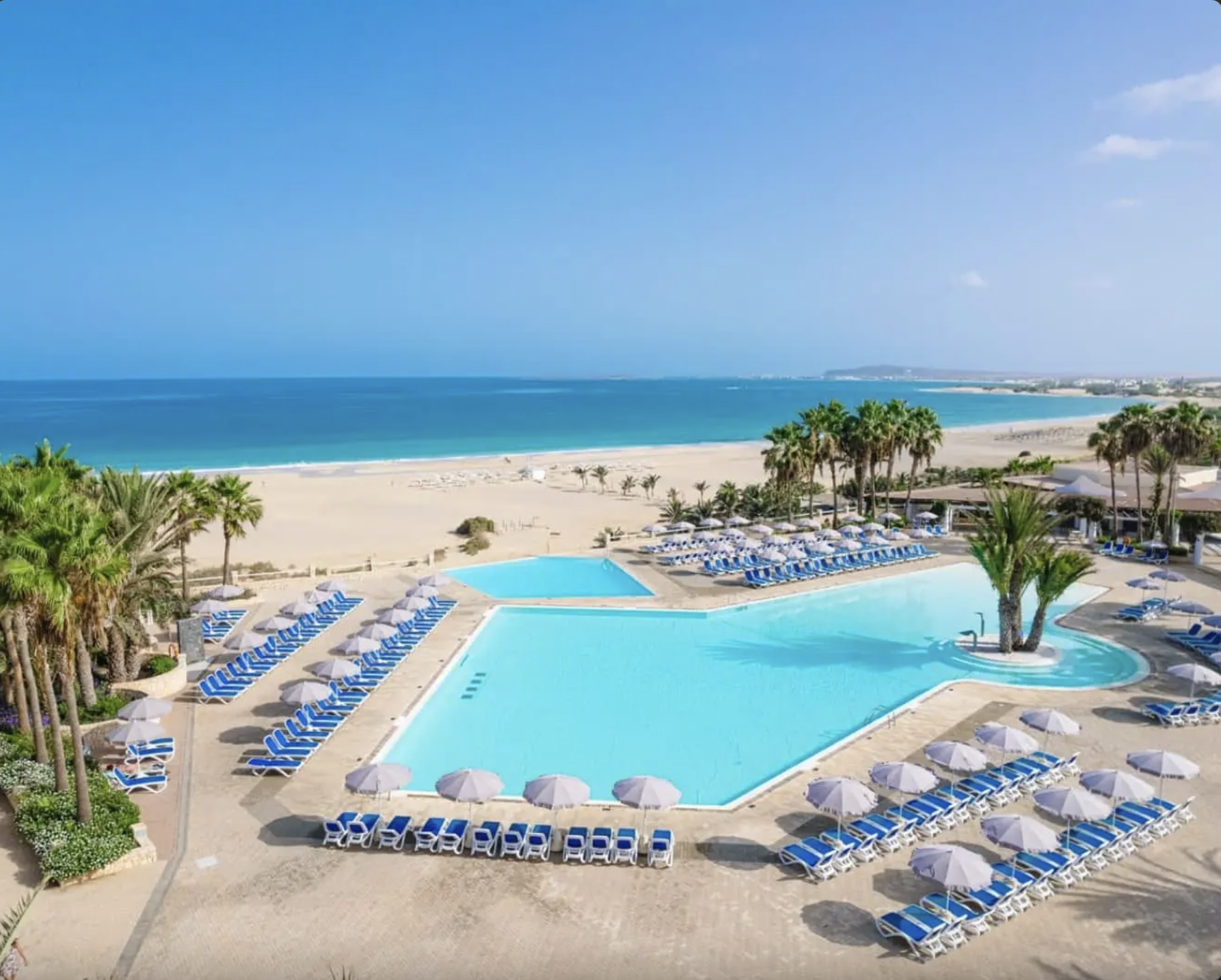 Piscina all'aperto con sedie a sdraio e ombrelloni, palme e spiaggia con mare in background, cielo sereno.