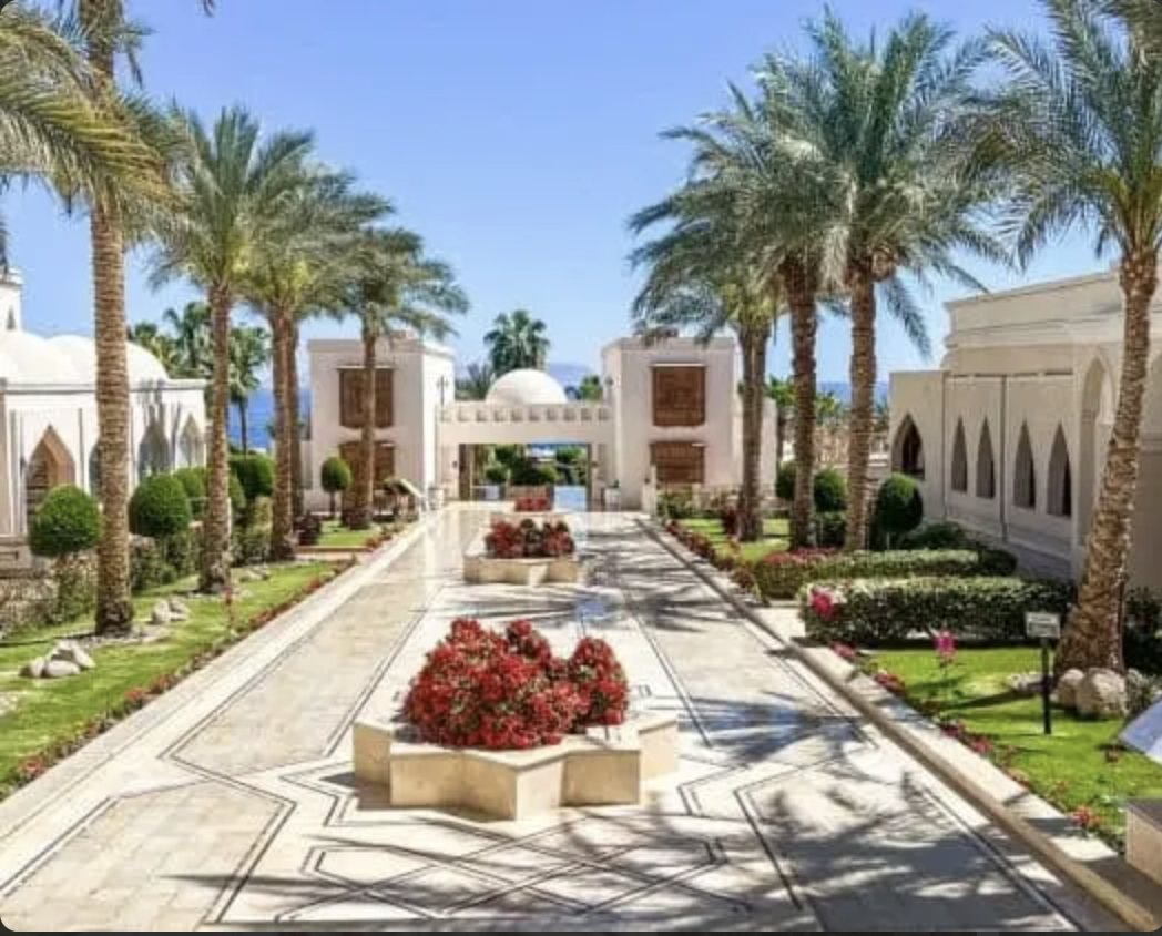 Giardino tropicale con palme, aiuole di fiori colorati e architettura in stile arabo su un percorso di marmo sotto un cielo azzurro