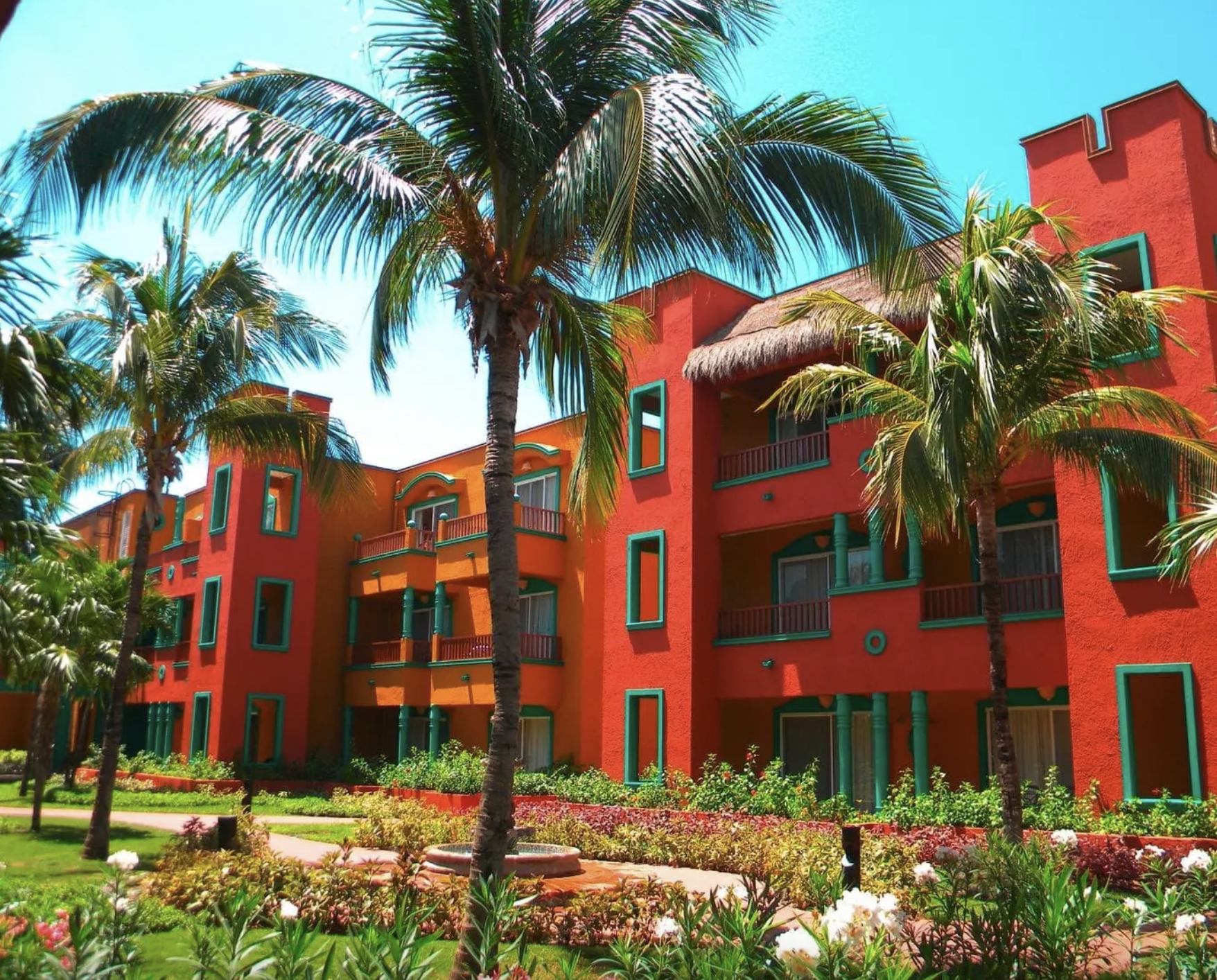 Edificio colorato con mura rosse, balconi, e finestre con cornici verdi, circondato da palme e giardino fiorito, sotto un cielo azzurro.