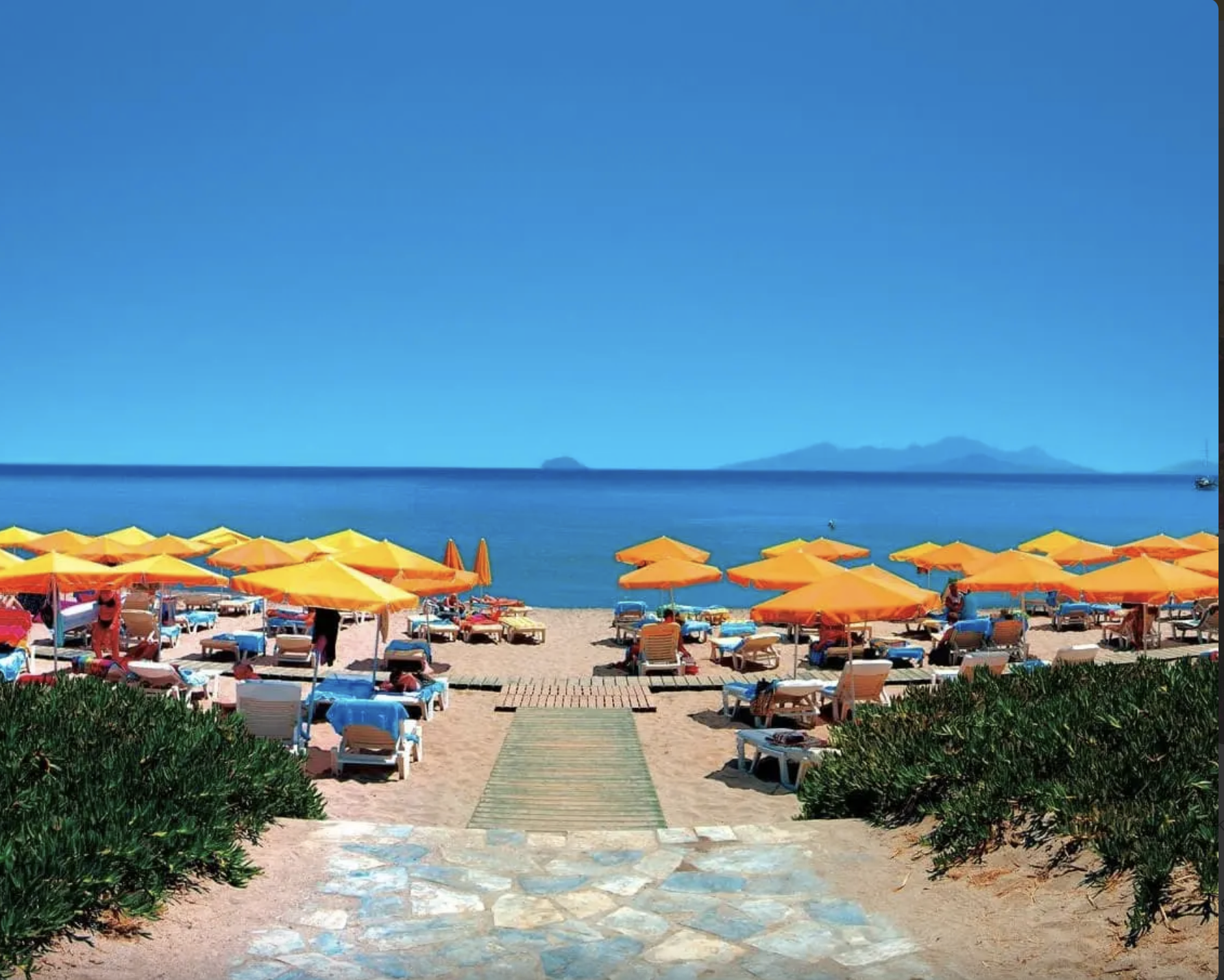 Spiaggia con ombrelloni arancio e gialli, lettini e una passerella di legno che conduce al mare, con acque azzurre e montagne sullo sfondo.