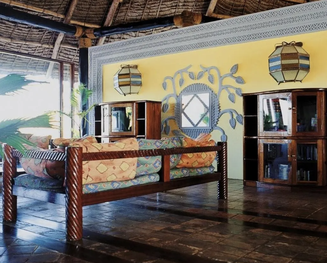 Interno di soggiorno con soffitto di paglia, parete gialla decorata con specchio e ramo con foglie, divano con coperte colorate, due armadi di legno in stile rustico e lampade colorate appese al soffitto.