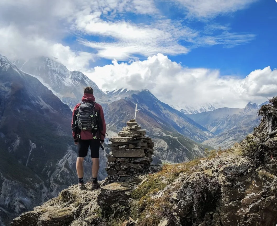 Escursionista in montagna con zaino, che guarda paesaggio alpino con montagne innevate, cielo nuvoloso e un accumulo di pietre segnato con un bastone.