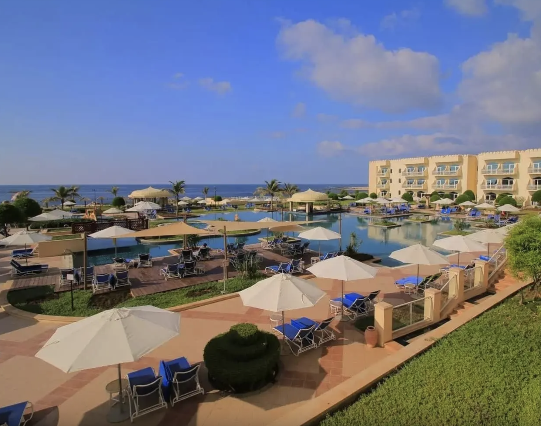Vista di una piscina di un resort sulla spiaggia con lettini, ombrelloni e palme, sullo sfondo il mare e un edificio di quattro piani.