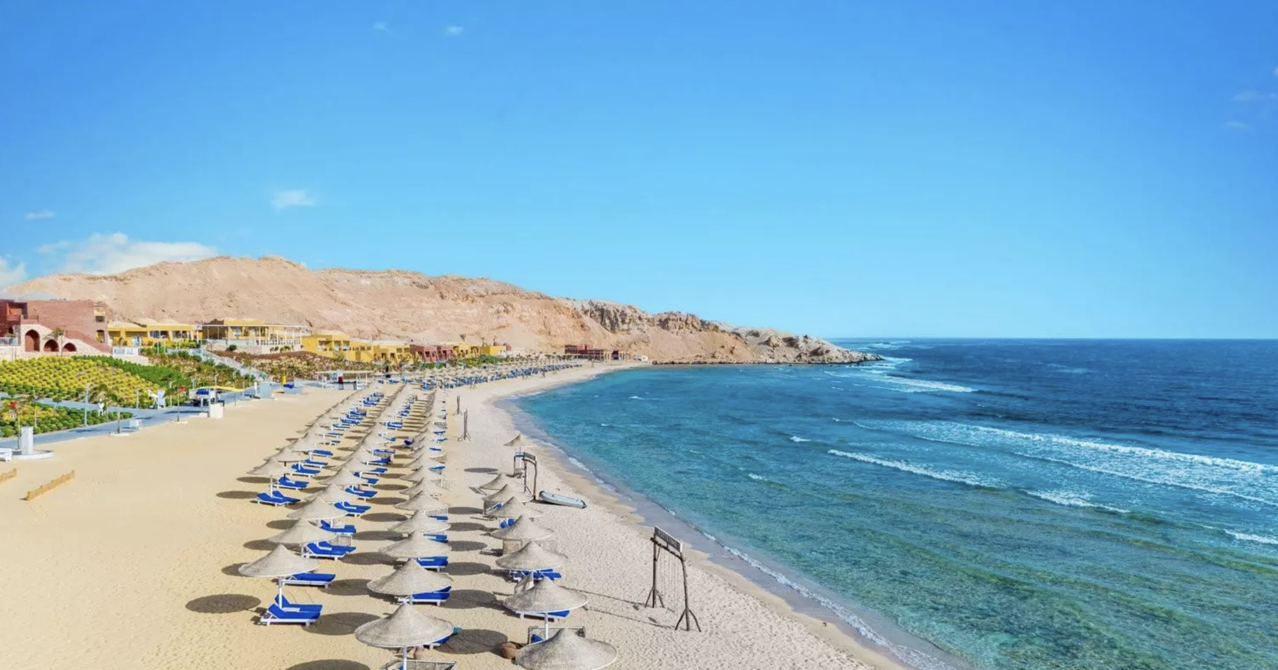 Spiaggia di sabbia con file di ombrelloni e sedie a sdraio, mare blu con piccole onde e colline sullo sfondo, cielo sereno.