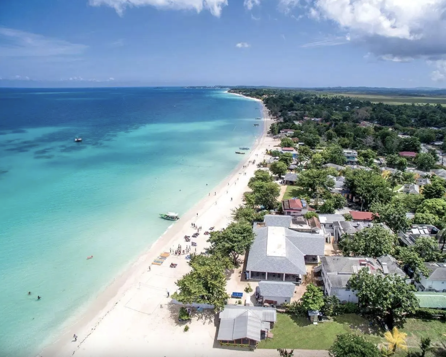 Spiaggia con acqua cristallina e palme, hotel e case lungo la costa, con persone e barche nel mare, sotto un cielo nuvoloso.