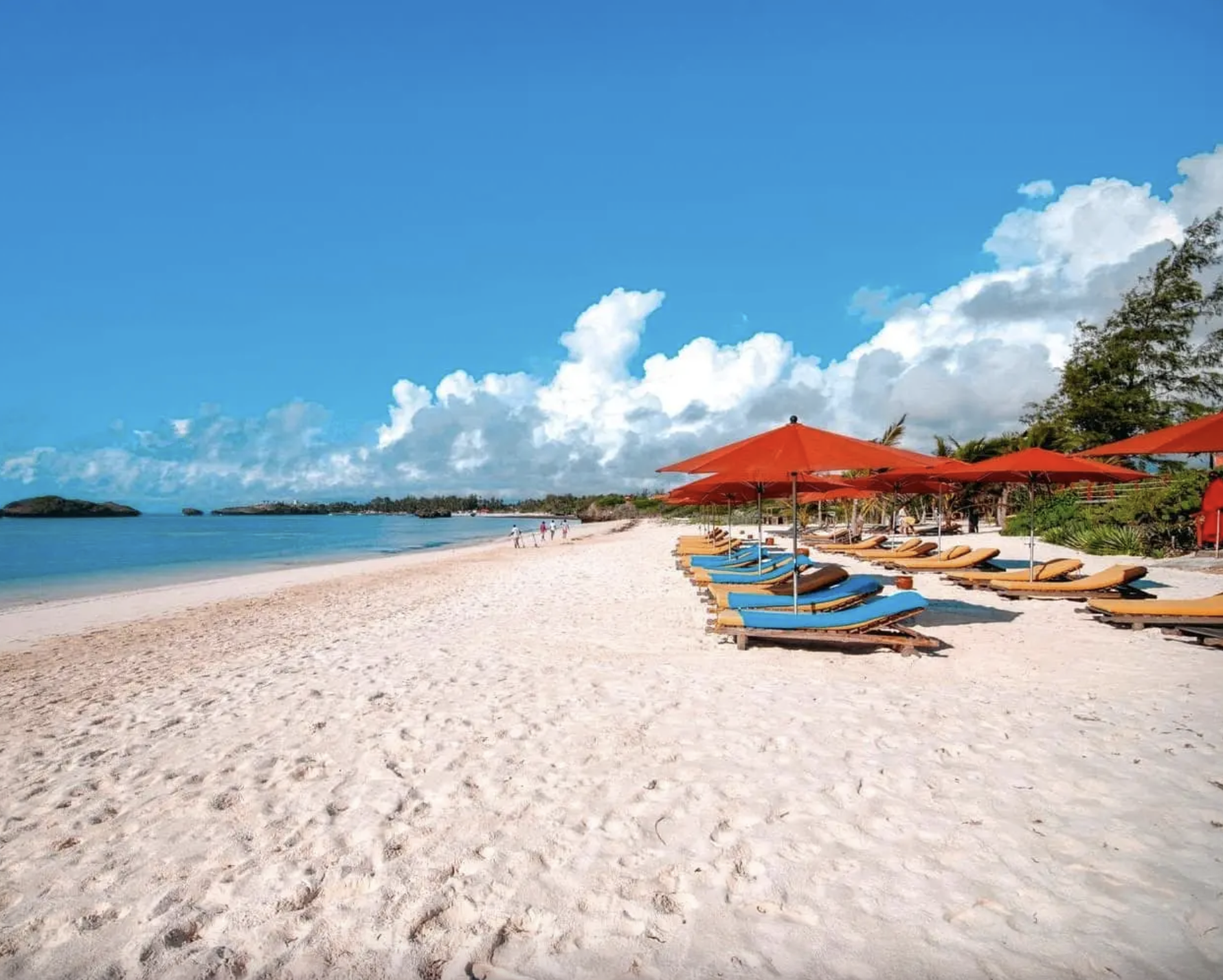 Spiaggia soleggiata con sedie a lettino e ombrelloni arancioni, mare blu e cielo con nuvole bianche, vegetazione verde sullo sfondo.