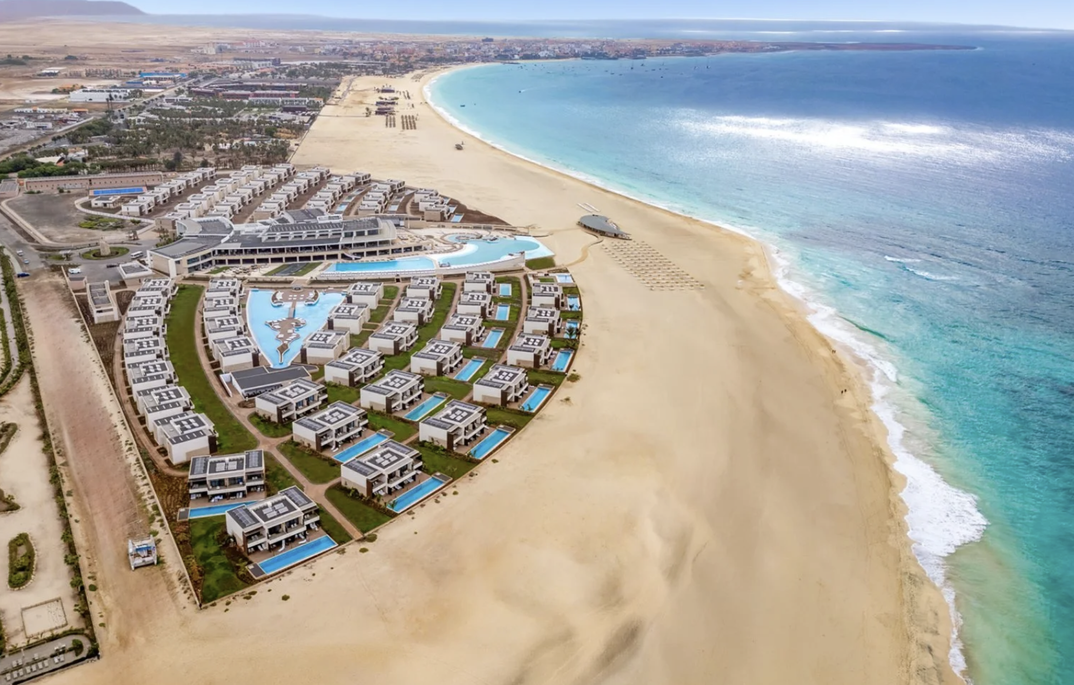 Vista aerea di un complesso turistico sulla spiaggia con ville, piscine e strutture moderne vicino al mare azzurro.