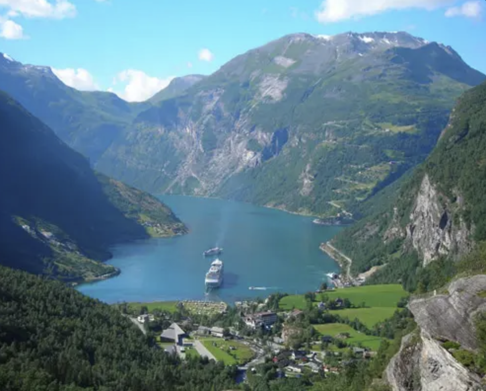 Paesaggio montano con un lago al centro, due navi e un paesino, circondato da montagne verdi e cielo sereno.