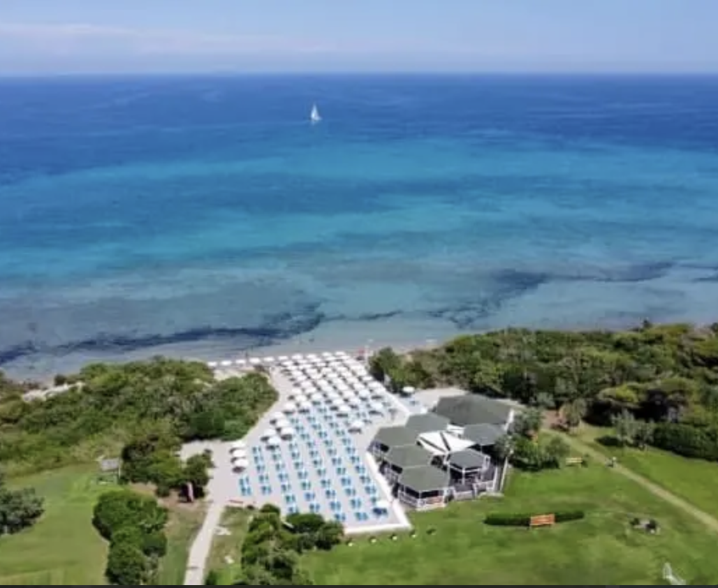 Spiaggia con lettini e ombrelloni, edificio vicino alla spiaggia, mare con nave in lontananza, vegetazione verde.