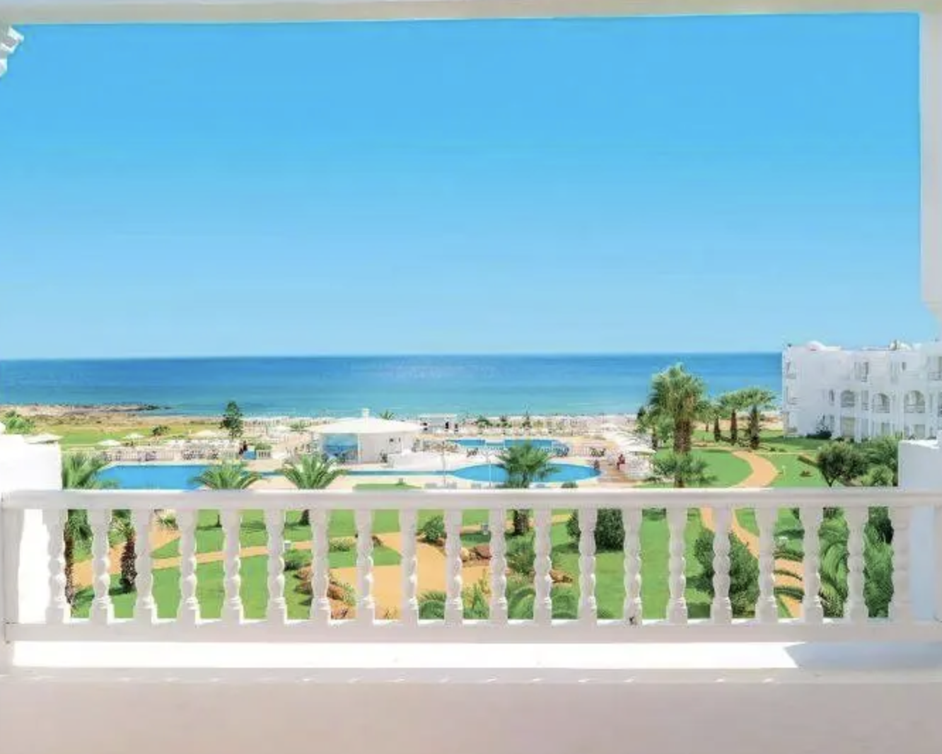 Vista panoramica di una spiaggia con mare blu, piscina, palme e edifici bianchi, vista da un balcone con parapetto bianco.
