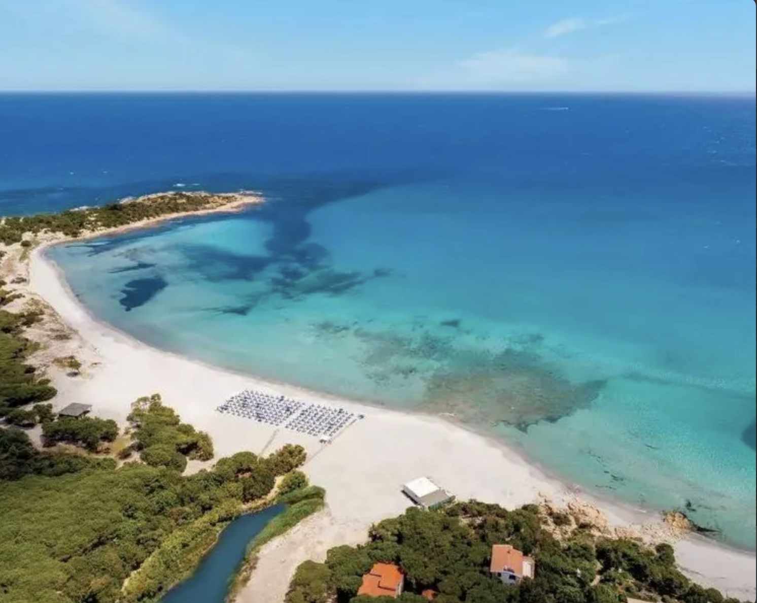 Spiaggia con acque cristalline, lettini e ombrelloni, verdeggianti dintorni e piccole strutture vicino alla costa.