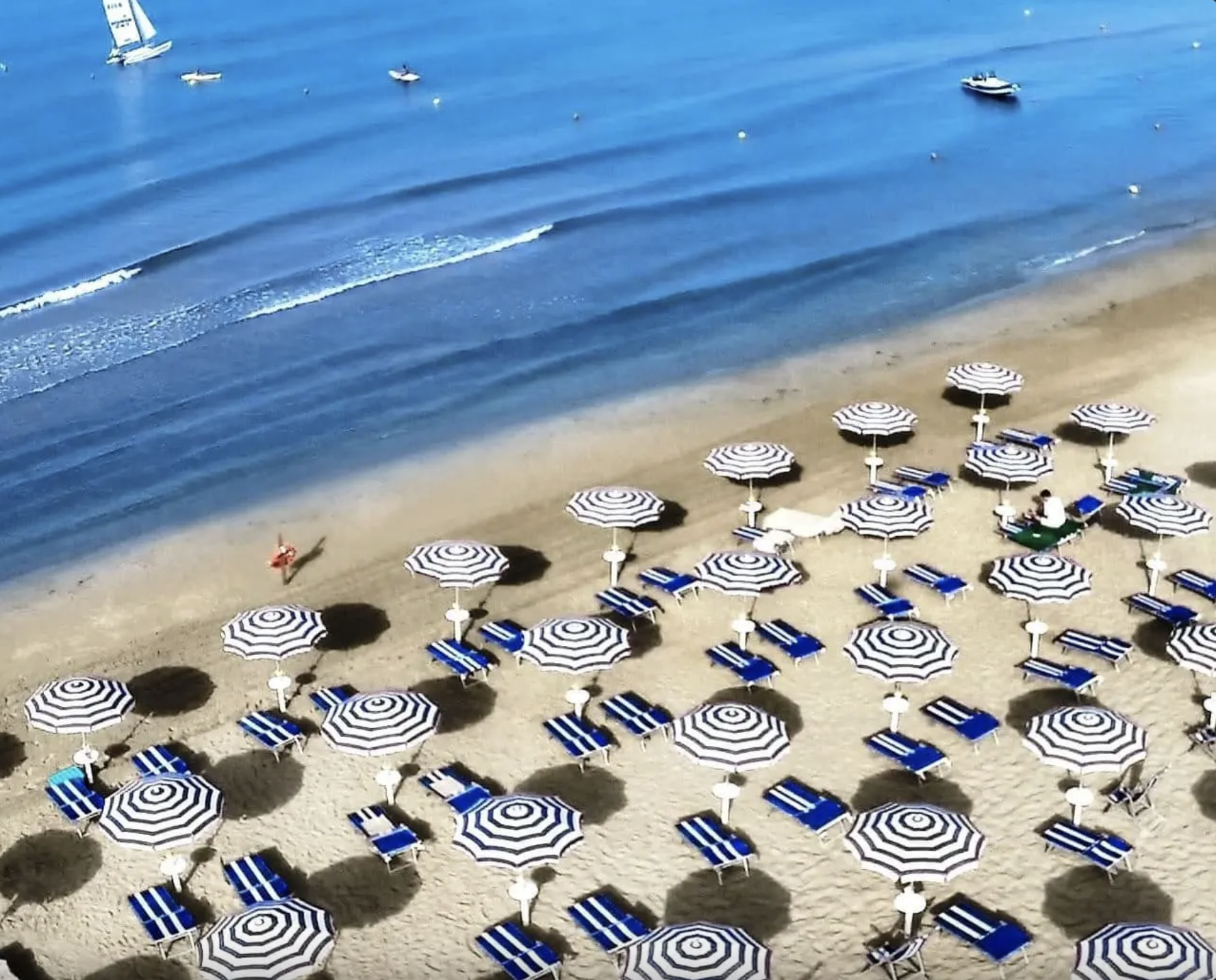 Spiaggia con ombrelloni a righe blu e bianche, lettini e team in spiaggia, vista dell'acqua con barche a vela e barche a motore.